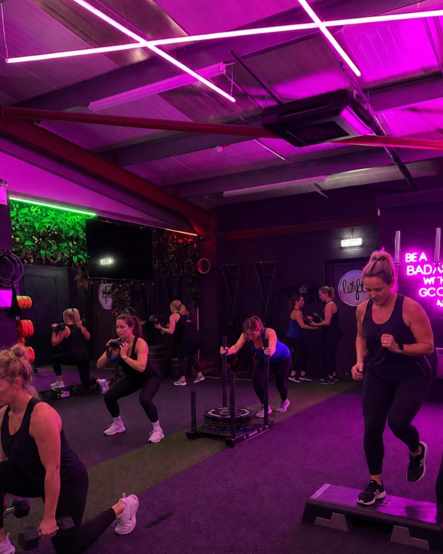 Women participating in a group fitness class in a gym with colorful neon lighting, including pink, green, and purple lights, engaging in various exercises such as step aerobics, boxing, and using equipment like weights and sleds.