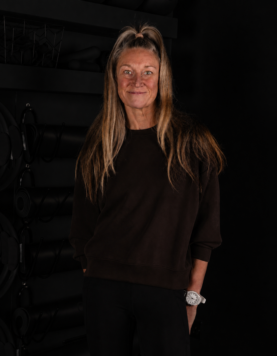 A woman standing in front of a black wall with shelves holding hats, with a neutral expression and hands in her pockets.