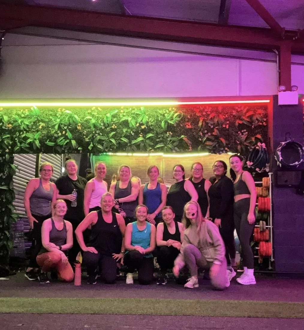 A group of 14 women and one man at a gym, posing for a photo together. They are in workout clothes, and the background features green plants, neon lighting in green, yellow, and red, and gym equipment.