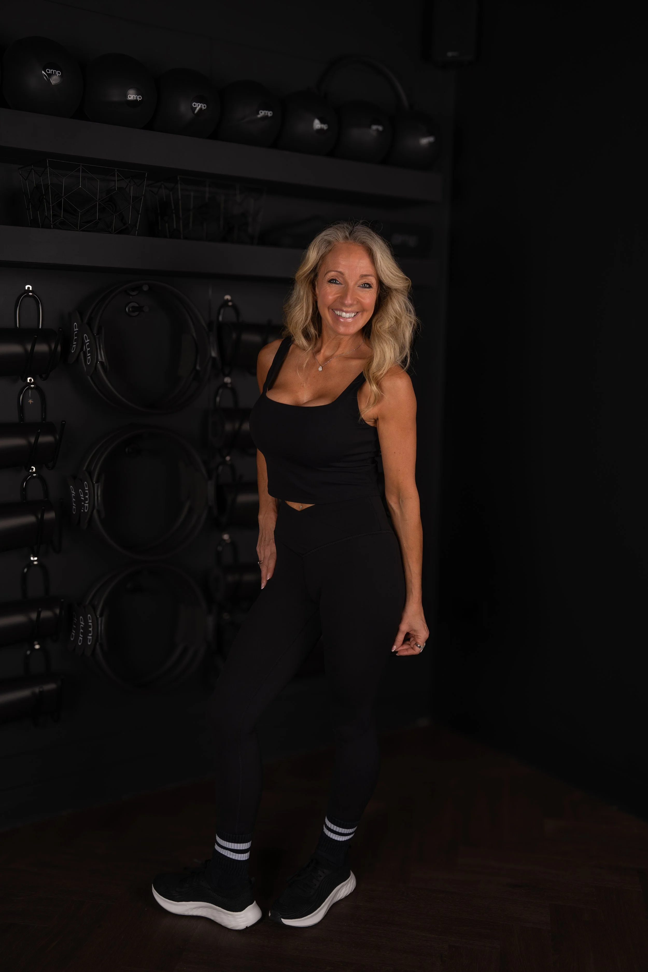 A woman with blonde hair wearing a black athletic top, black leggings, black and white striped socks, and black sneakers standing in front of a black wall with gym equipment including kettlebells, dumbbells, and resistance bands, smiling at the camera.