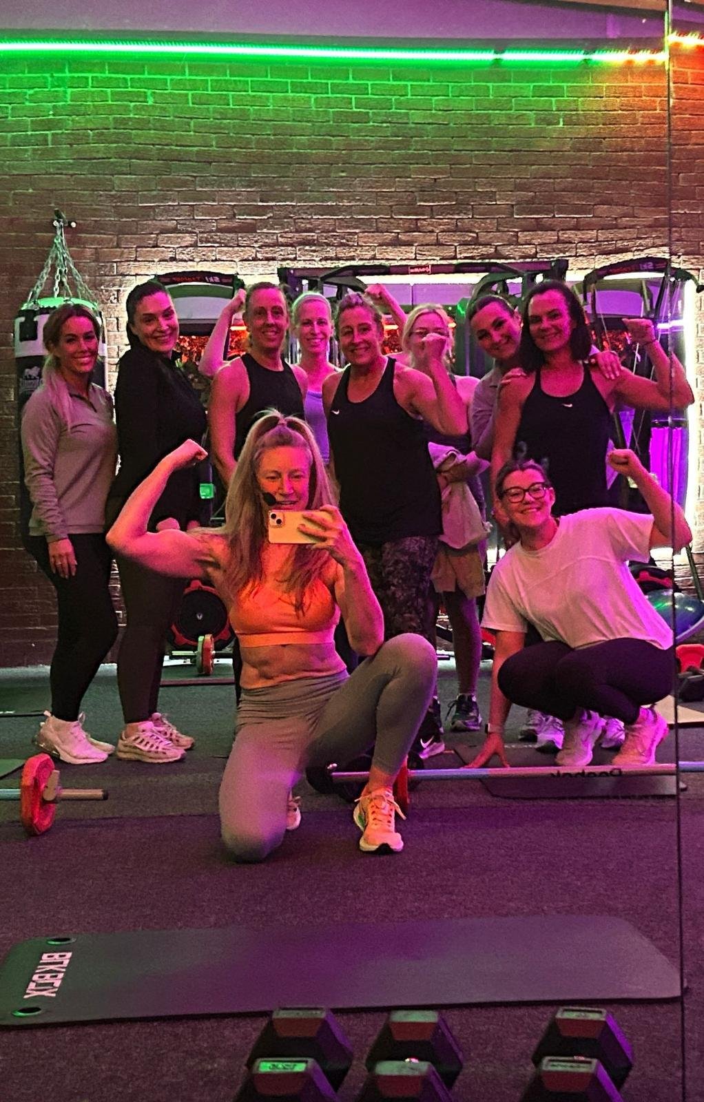 A group of women in workout attire posing for a photo in a gym with brick walls and neon lights.