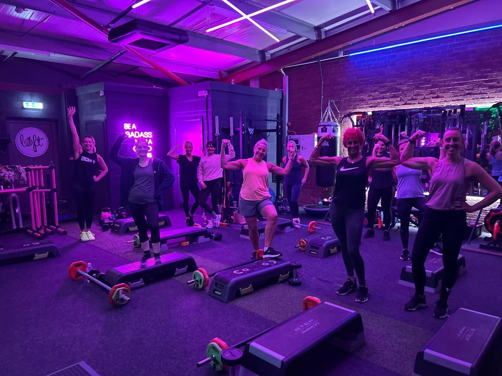 Group of women in a gym posing during a workout session under colorful neon lighting, with exercise equipment around them.