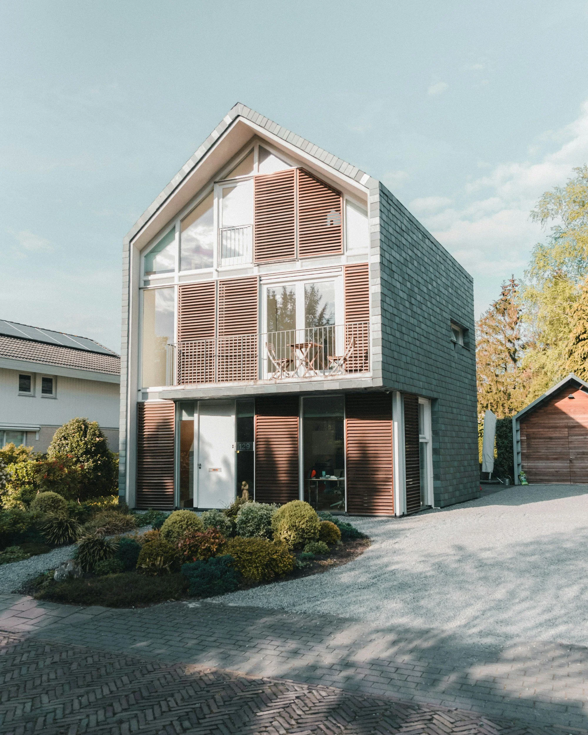 Modern house with large windows, wooden slats, small balcony, pitched roof with gray shingles, front garden with bushes, gravel driveway, and trees in the background.