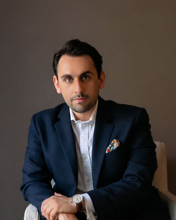 A man in a navy blazer and white shirt sitting against a gray background.