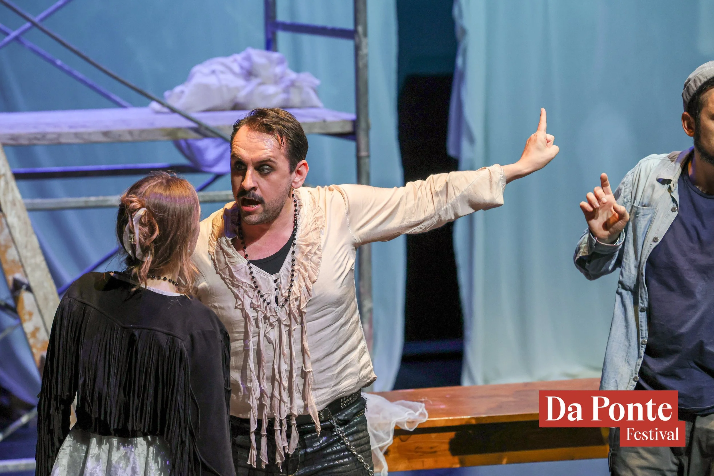 A scene from a theatrical performance with a man gesturing passionately towards a woman, with a scaffolding structure in the background and a sign that reads "Da Ponte Festival" in the bottom right corner.