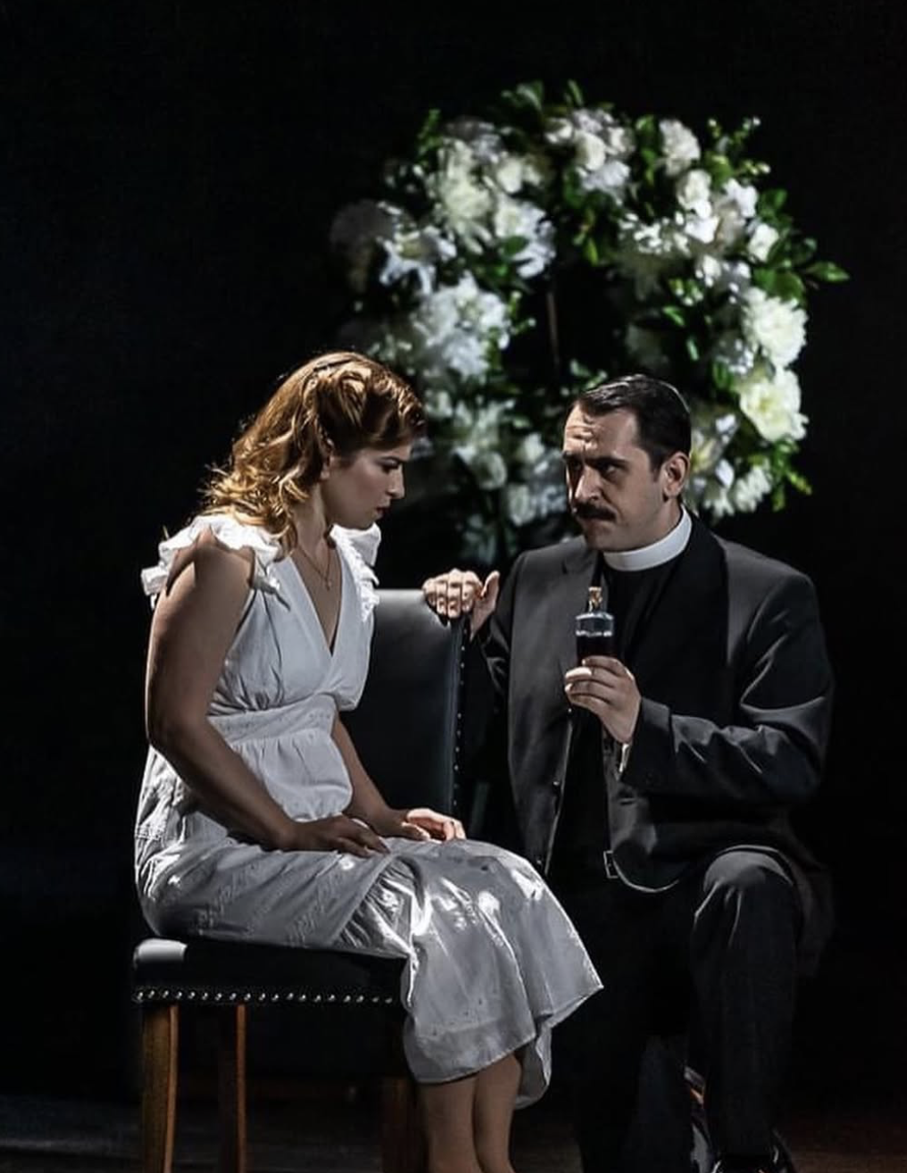 Conall O'Neill, Bass. Wexford Festival Opera. A scene from a stage play showing a woman in a white dress sitting on a chair, and a priest or religious figure kneeling beside her, holding a small bottle. A floral arrangement in the background.