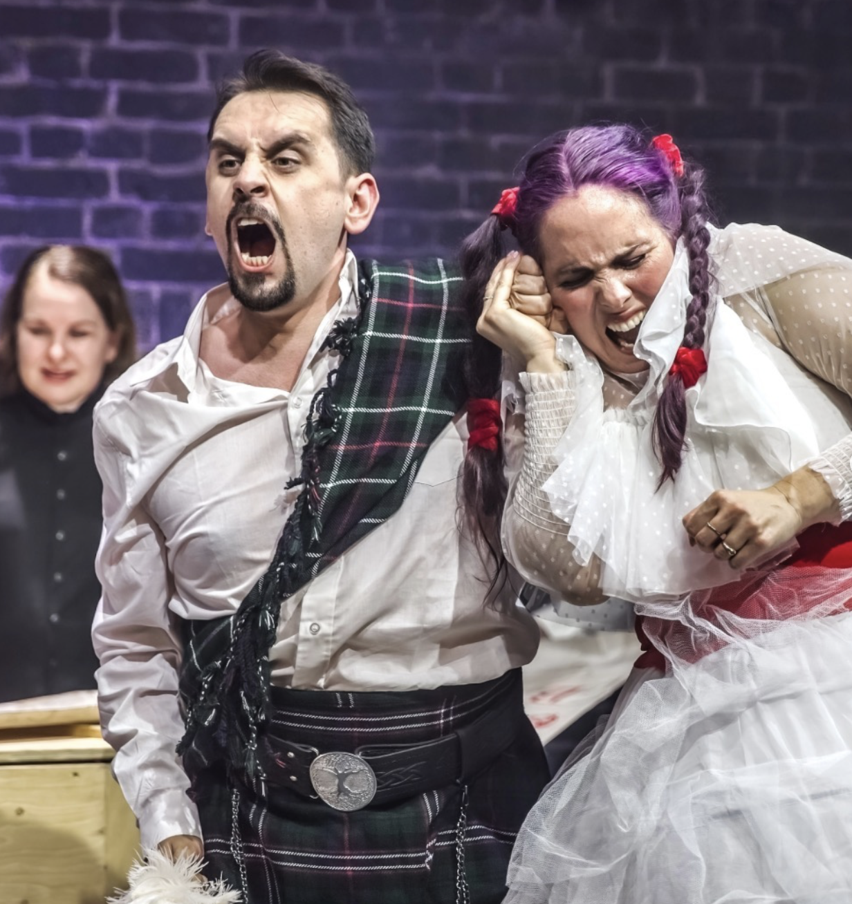 Three actors performing on stage, with the man on the left wearing a traditional Scottish kilt and white shirt, shouting or singing passionately. The woman on the right has purple hair styled in braids with red ribbons, wearing a white dress with sheer polka-dot sleeves and ruffles, and appears to be distress or crying, with her hand on her ear. The background is a dark brick wall, and another performer can be seen in the background.