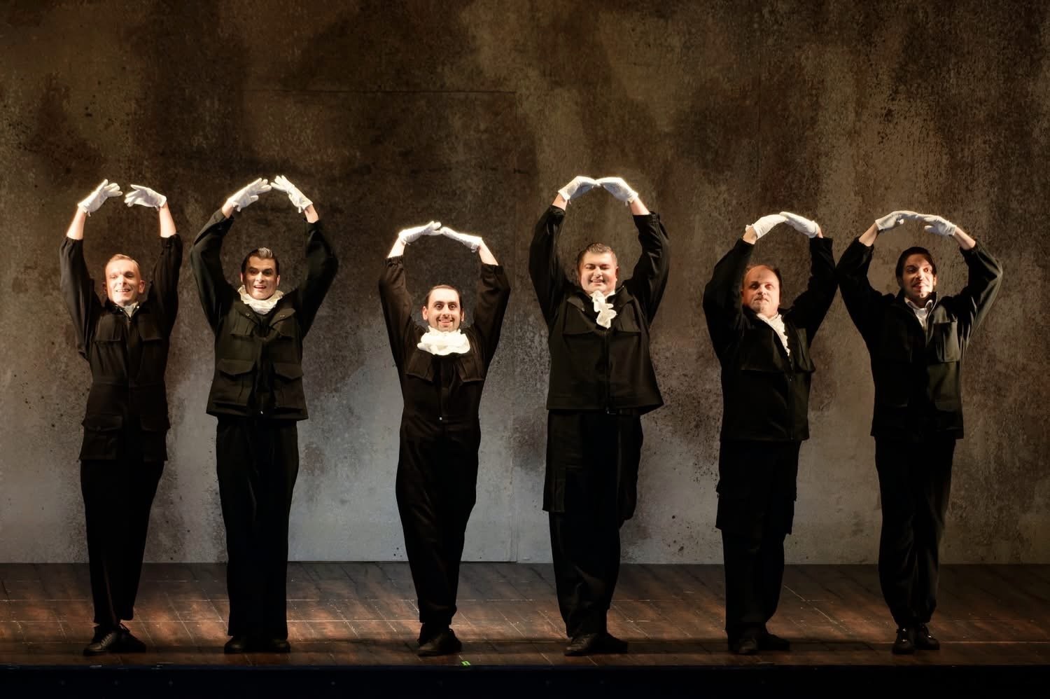 Conall O'Neill, Bass. A Midsummer Night's Dream, Lithuanian National Opera. Six men dressed in black costumes and white gloves standing on a stage with a plain wall background, performing a synchronized dance move with their arms curved overhead.