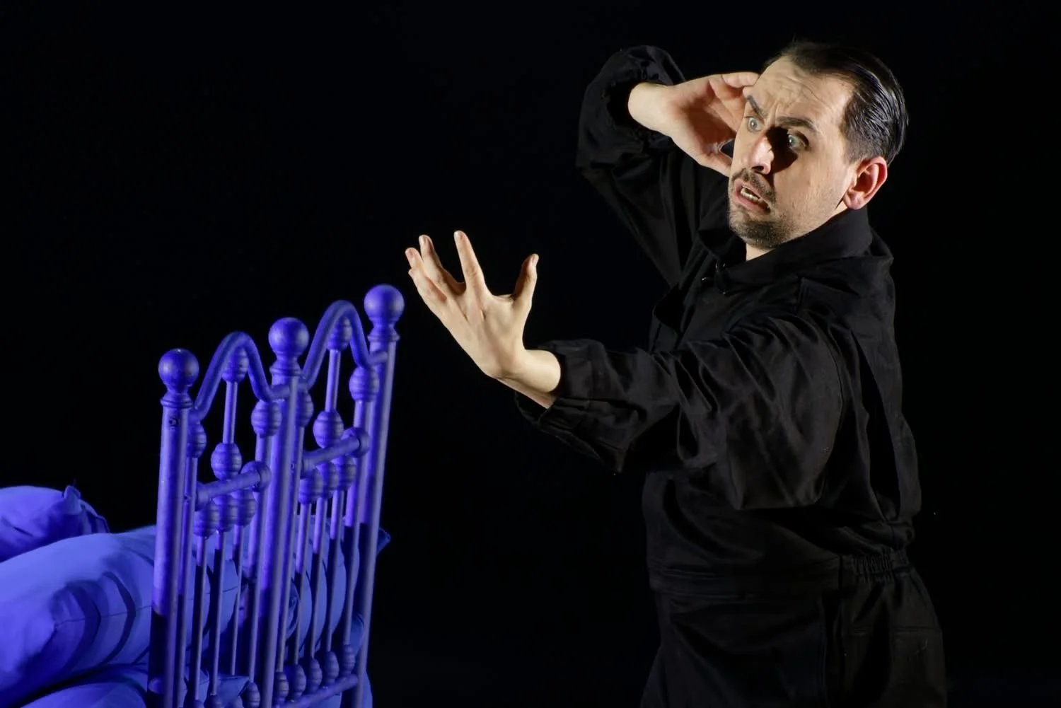 Conall O'Neill, Bass. A Midsummer Night's Dream, Lithuanian National Opera. A man with dark, slicked-back hair and a beard, wearing black, gesturing with his hands toward a bed with a purple headboard against a black background.