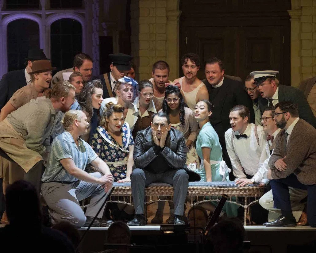 Conall O'Neill, Bass. Sir John in Love, BYO/Opera Holland Park. A group of theater actors on stage, with one man sitting center in leather jacket, surrounded by other actors in period costumes, some smiling, some with surprised expressions.