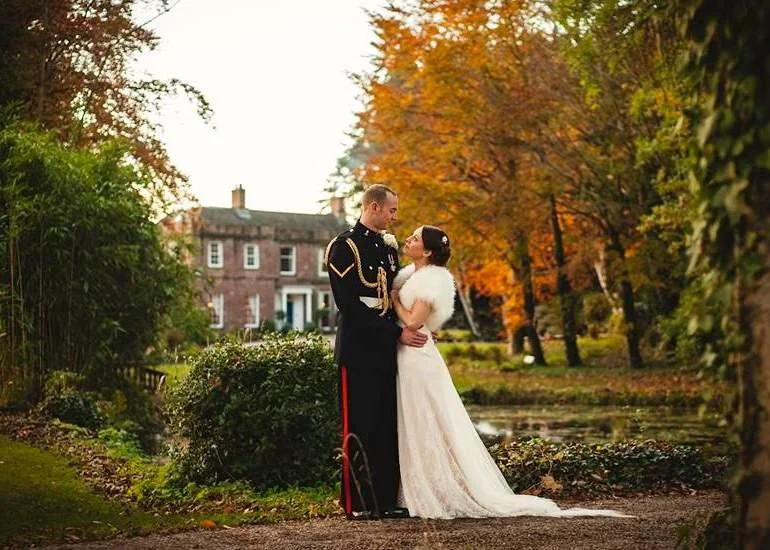 A bride and groom in wedding attire standing close together in a park-like setting during autumn, with trees showing fall colors and a large house in the background.