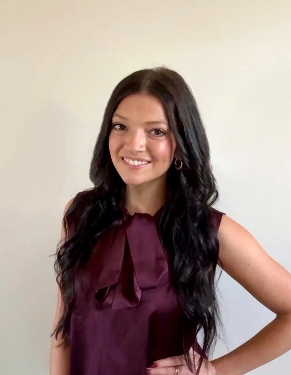A young woman with long, wavy dark hair, smiling and wearing a sleeveless maroon top with a bow tie at the neck, standing against a plain light-colored wall.