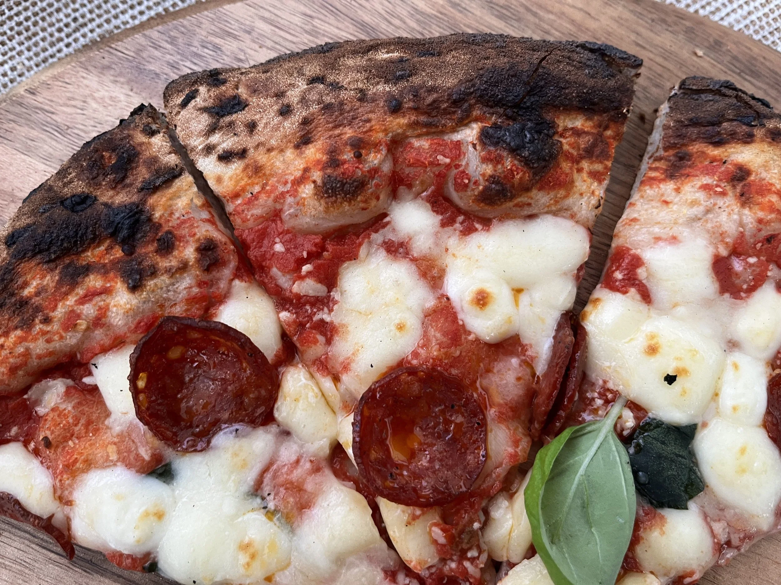 Close-up of a homemade pepperoni and cheese pizza with a slightly burned crust on a wooden surface, garnished with a basil leaf.