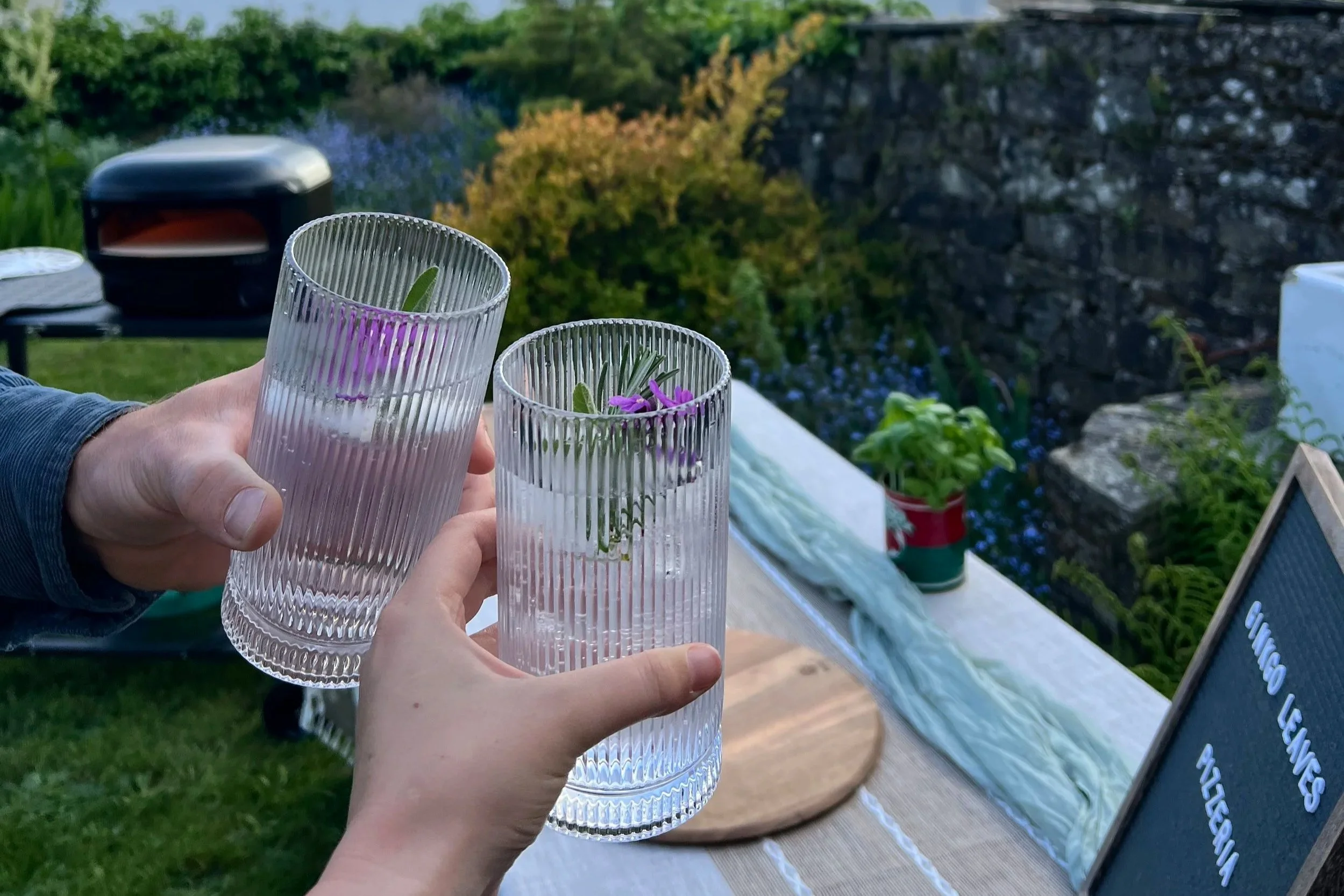 People raising glasses with flowers in an outdoor setting, with a garden, stone wall, and a sign in the background.