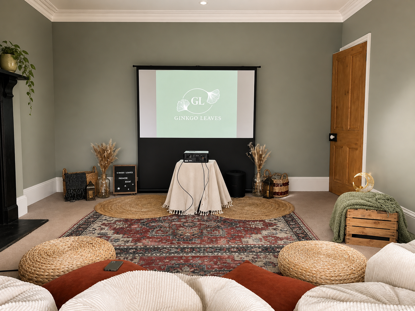 Living room set up for a presentation with a projector on a small table, a large screen displaying 'Ginkgo Leaves' logo, decorative baskets, vases with dried plants, and cozy seating with poufs and rugs.