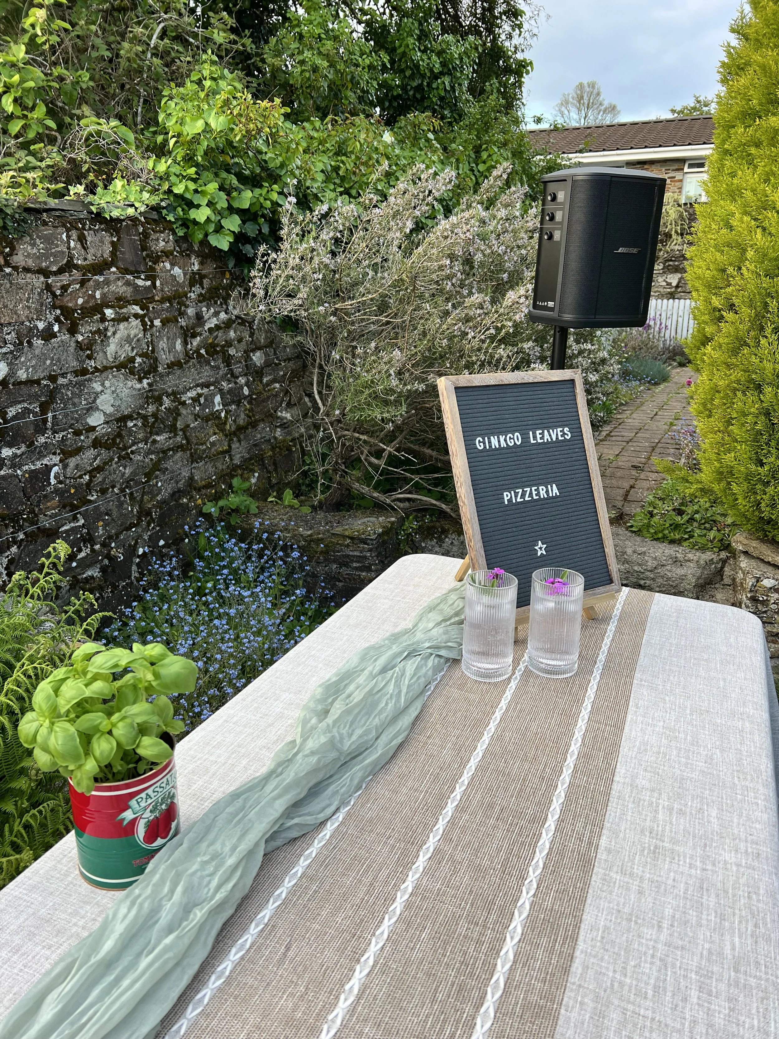 An outdoor dining setup with a table covered with a beige tablecloth and a green cloth runner. On the table, there are two glasses with pink flowers inside. Behind the table, there is a black speaker on a stand, a signboard that reads "Ginkgo Leaves Pizzeria," a tin can with a basil plant, and a garden with various green plants and flowers, including a bush with white flowers, a yellow-green hedge, and small blue flowers. A brick pathway runs through the garden, and a house with a tiled roof is visible in the background under a partly cloudy sky.