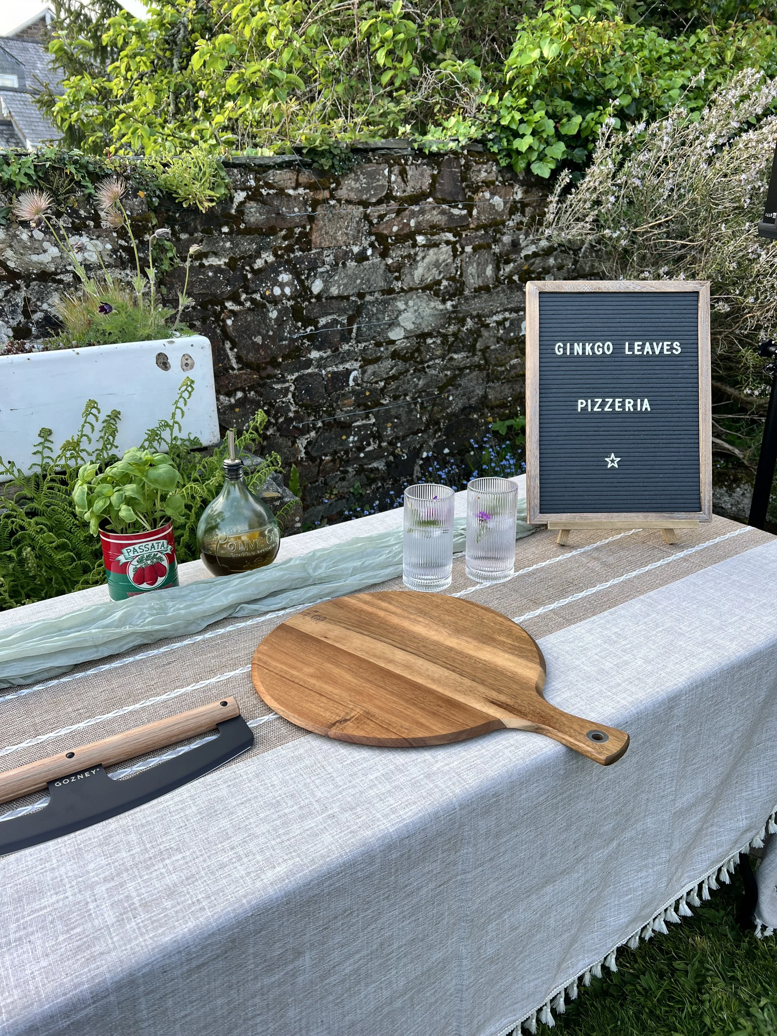 Outdoor dining table with a wooden cutting board, a pizza cutter, several glasses of water, a potted herb plant, a bottle of olive oil, and a sign that reads 'Ginkgo Leaves Pizzeria' against a stone wall and greenery background.