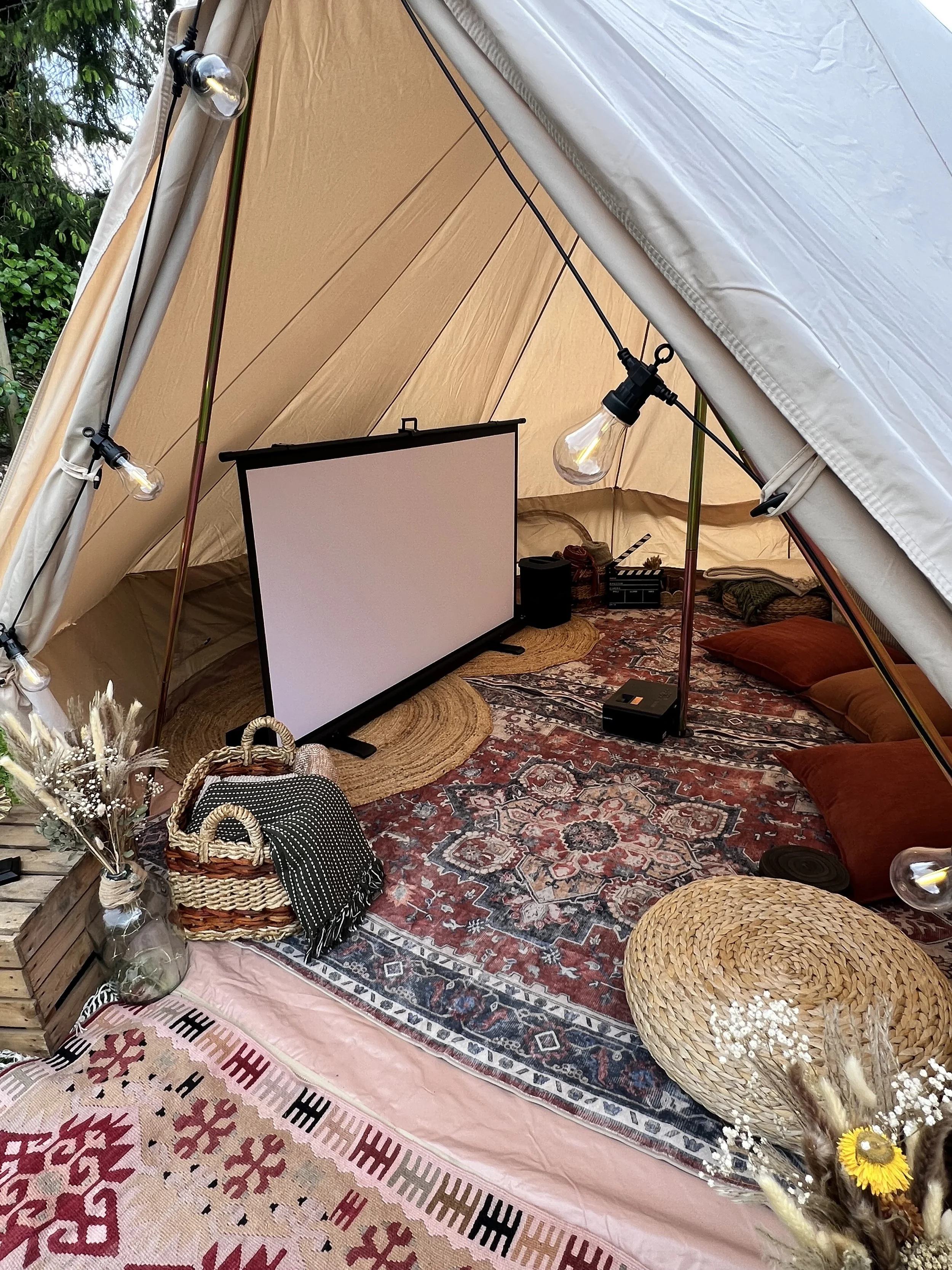 A cozy indoor space with a tent, decorated with string lights and vintage style lighting bulbs. There is a large screen, a patterned rug, and cushions on the floor. Decor items include wicker baskets, dried flowers, and a variety of textiles arranged to create a warm, inviting atmosphere.