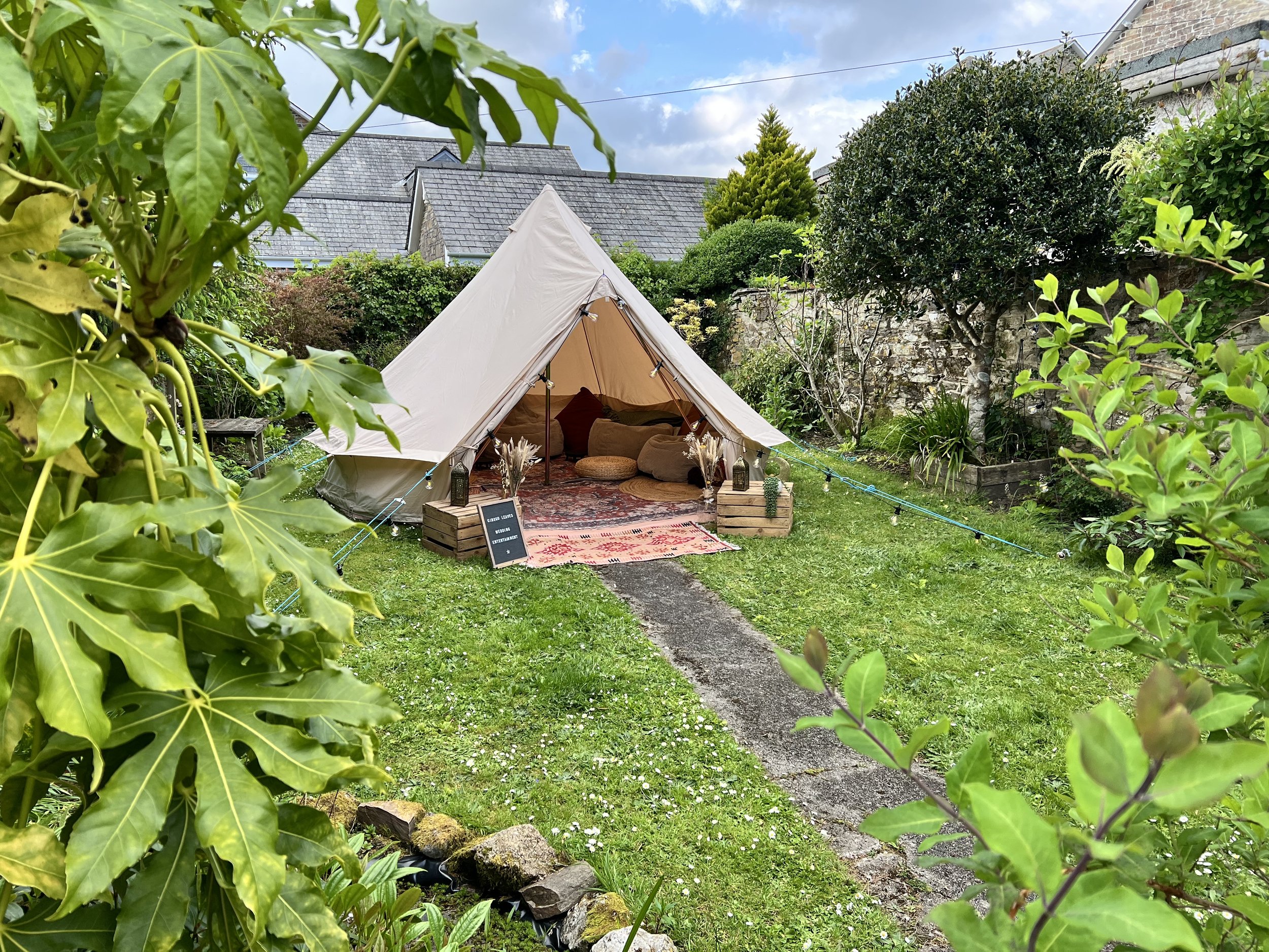 A view of a backyard garden with a white canvas teepee tent featuring cushions and a rug inside. The garden is lush with green plants, trees, and a stone wall in the background. String lights are hung along the tent, creating a cozy ambiance.