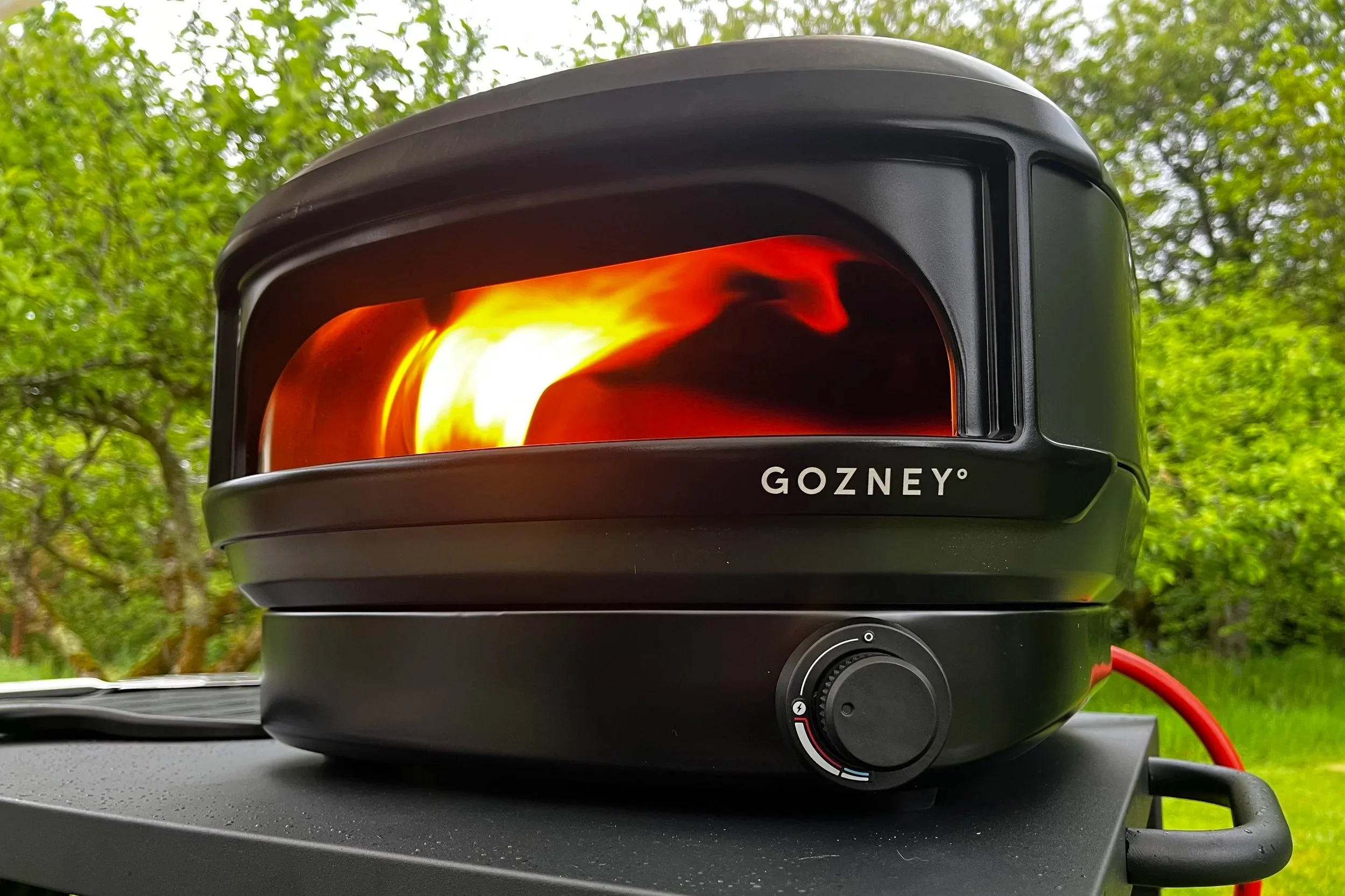 A black portable barbecue smoker with a glowing orange and yellow fire inside, placed on a table outdoors, with blurred green trees in the background. The smoker has a round control knob with temperature settings and the brand name 'GOZNEY' on the front.