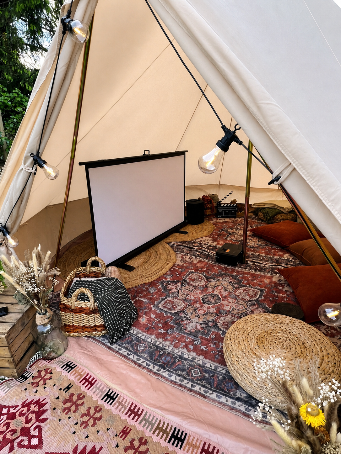 A cozy outdoor tent setup with a projector, a large white screen, and string lights. The space is decorated with patterned rugs, cushions, baskets, and dried flowers.