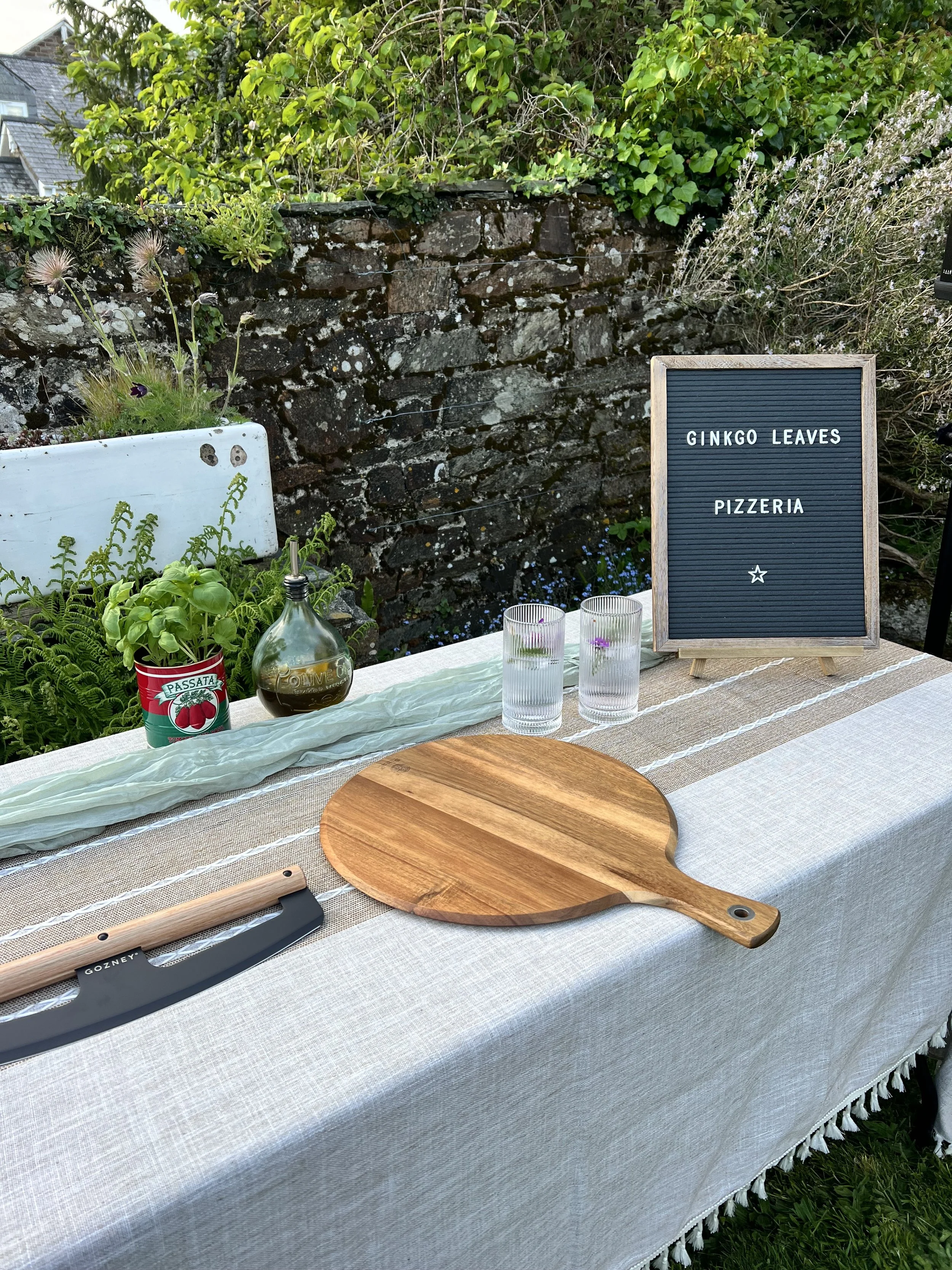 A table decorated with a light-colored tablecloth, a wooden cutting board, a set of knives, a potted herb plant, a bottle of olive oil, a chalkboard sign with the text "Ginkgo Leaves Pizzeria" and two drinking glasses with flowers inside, set outdoors against a stone wall and greenery.
