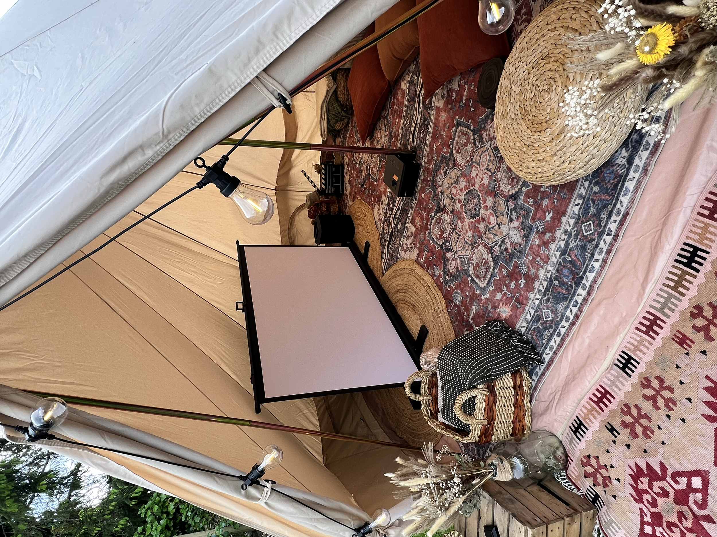 A cozy indoor space with a woven circular mat and small bouquet of flowers on a Persian rug, a wicker basket, and a folded blanket with a pink and white geometric pattern. There is a screen and some cushions on a low wooden platform, with string ligh