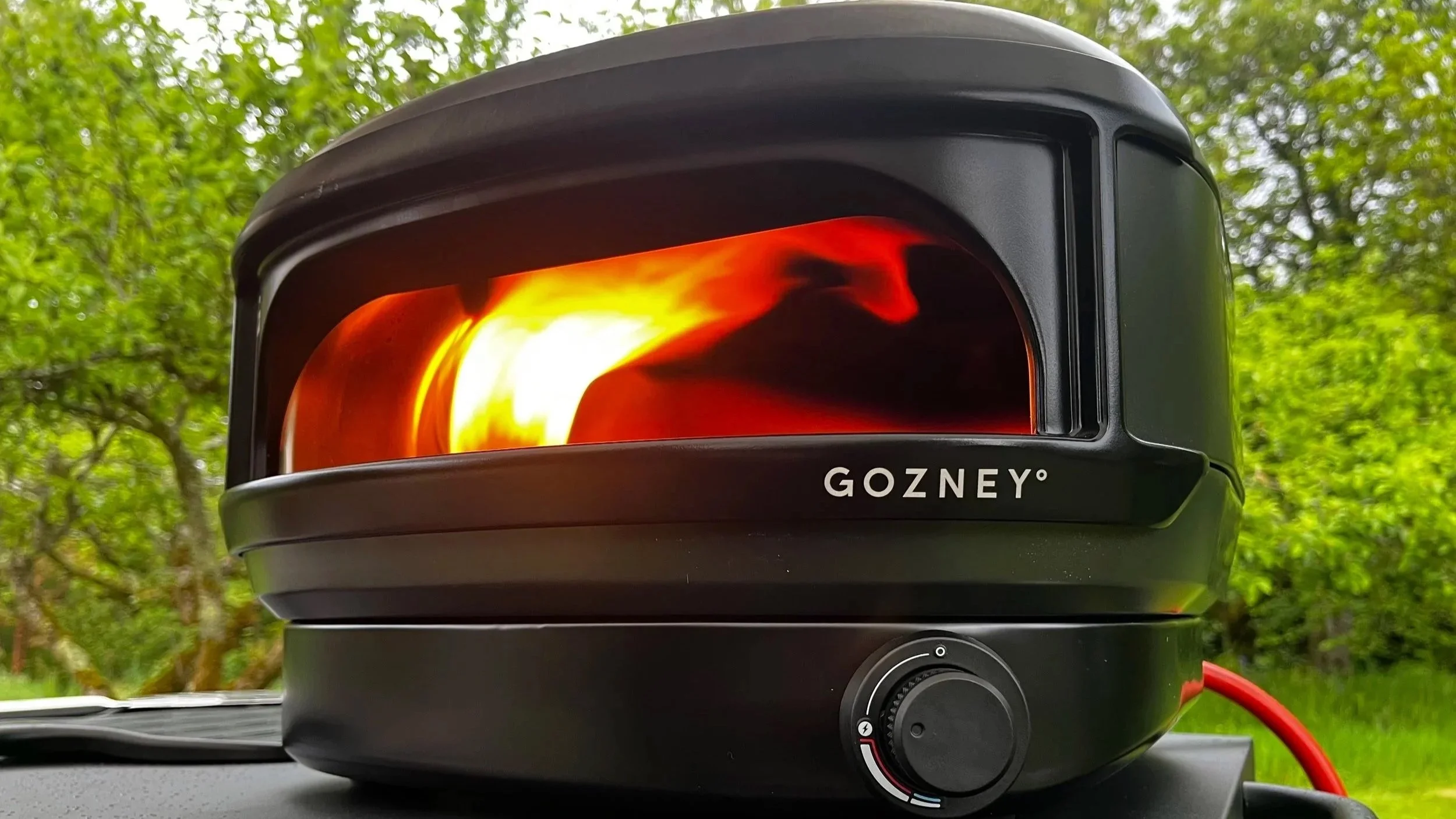 Gozney portable pizza oven with flame inside, outdoors with green trees in background.