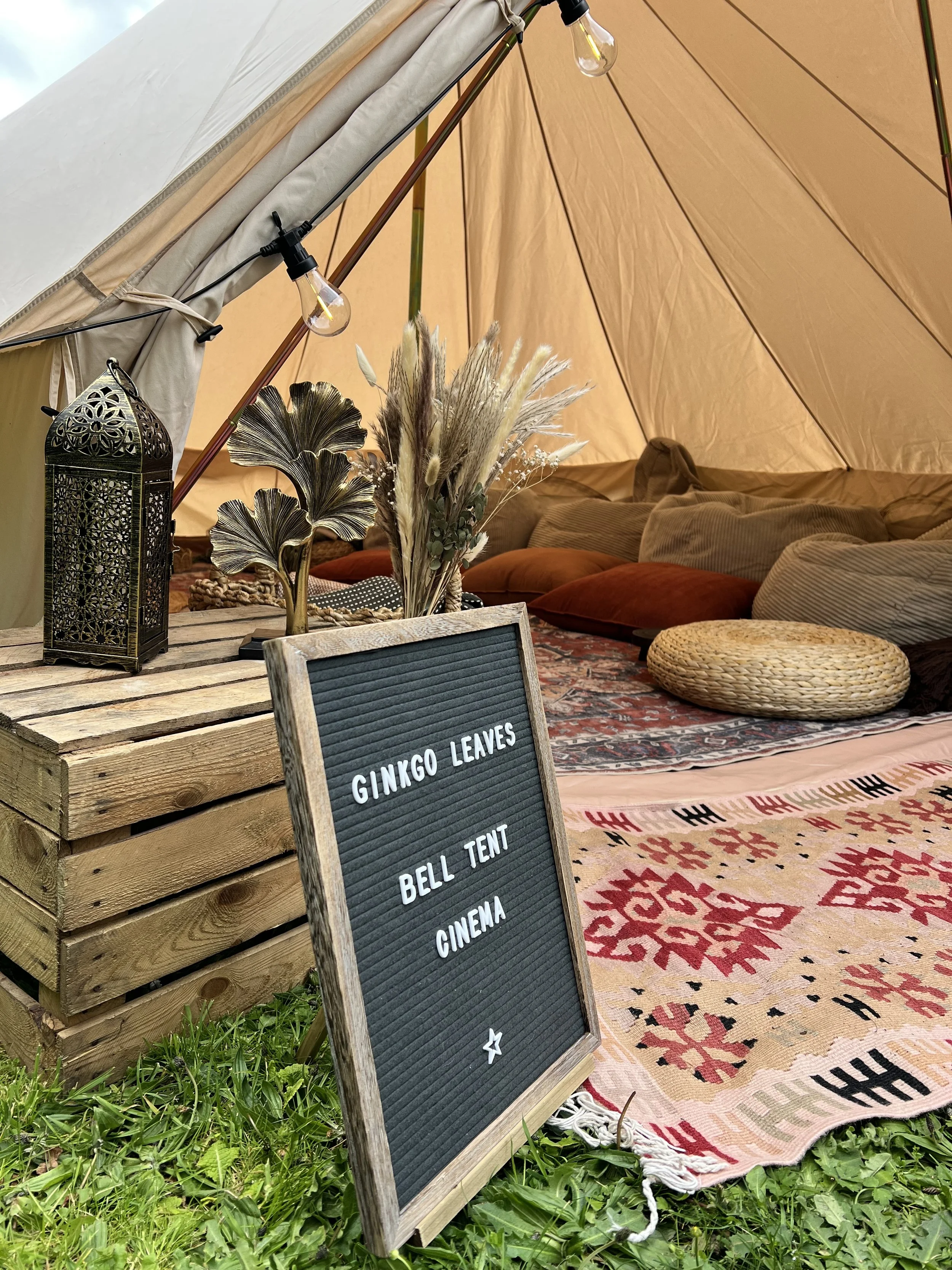 A cozy lounge area inside a tent with beige walls, decorated with dried foliage, a patterned rug on the floor, and cushions. There is a wooden table with a decorative lantern, a signboard with text, and string lights hanging above.