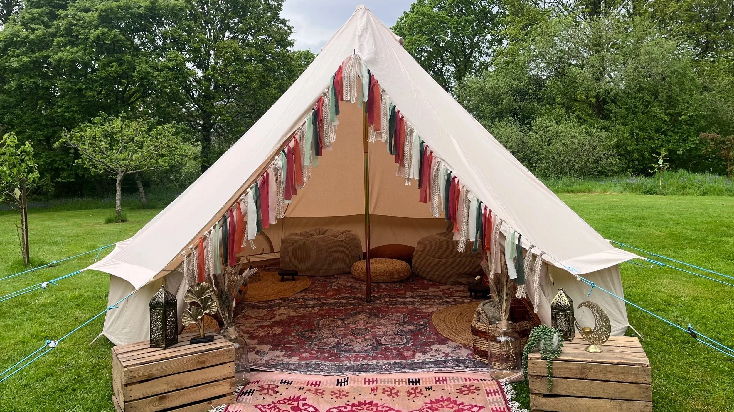 A large white camping tent set up on a grassy area surrounded by green trees. The tent is decorated with hanging fabric strips in various colors and patterns. Inside the tent, there are cushions, a rug, and decorative items such as lanterns and plants. The entrance is open, revealing a cozy interior with seating.