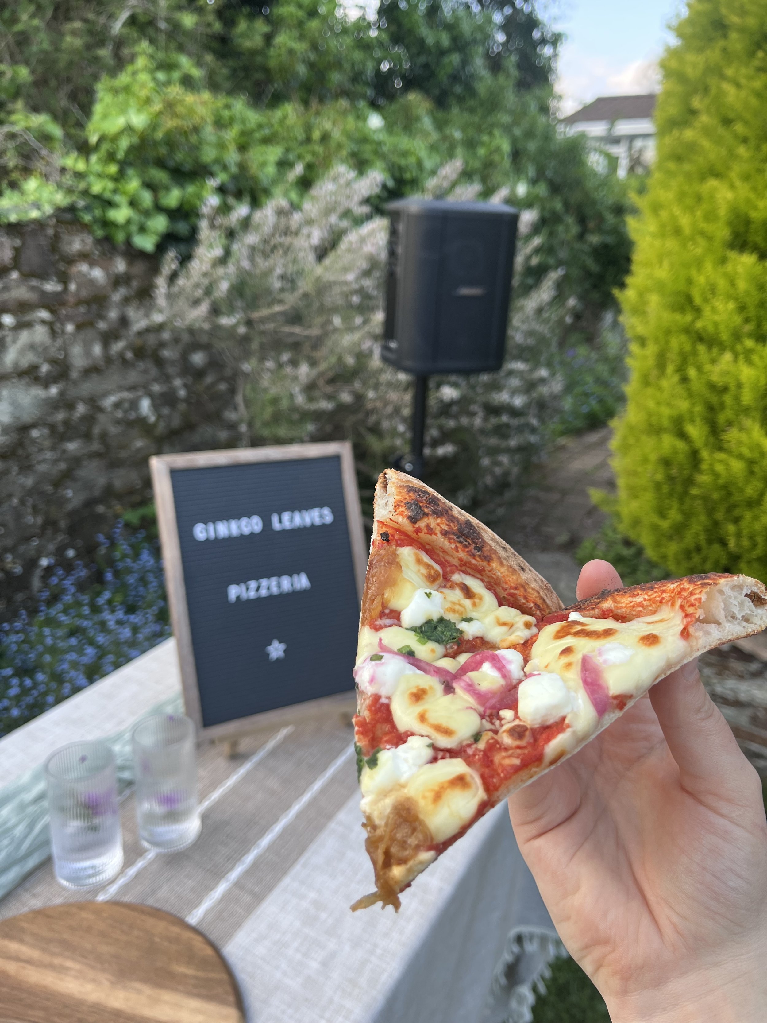 Close-up of a hand holding a slice of pizza with melted cheese, pepperoni, and vegetables. In the background, there is an outdoor setting with a black speaker, a sign that says "Ginkgo Leaves Pizzeria," two glasses with drinks, and green trees and bushes.