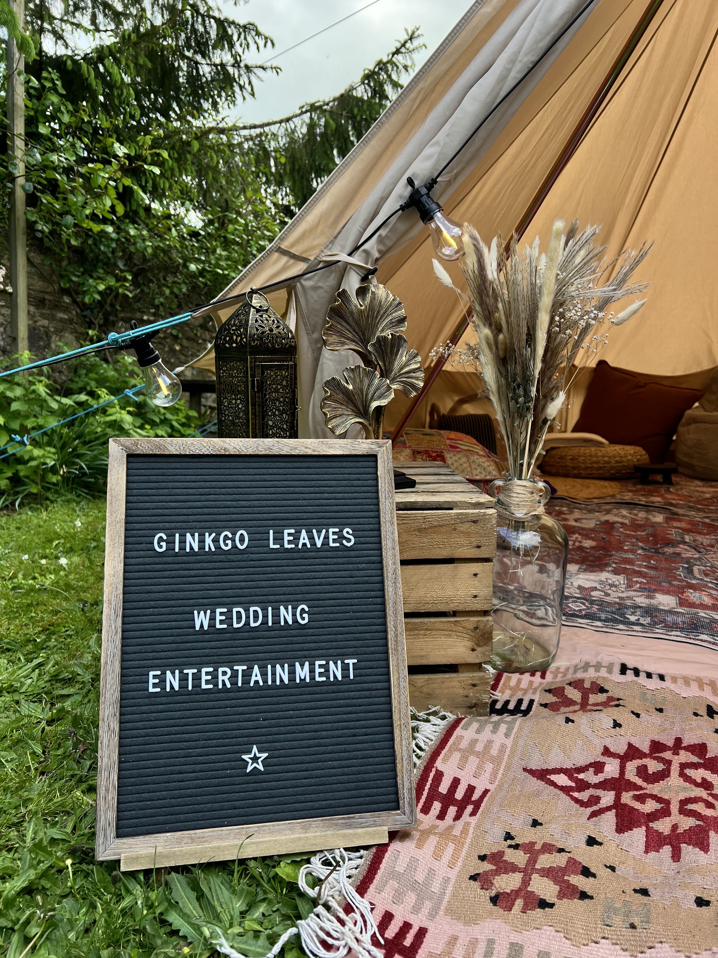 A decorative outdoor setting with a black letter board sign on a wooden crate, displaying words related to a wedding and entertainment. Behind the sign, there is a vase with dried flowers, a patterned rug, and a tent with string lights, surrounded by greenery.