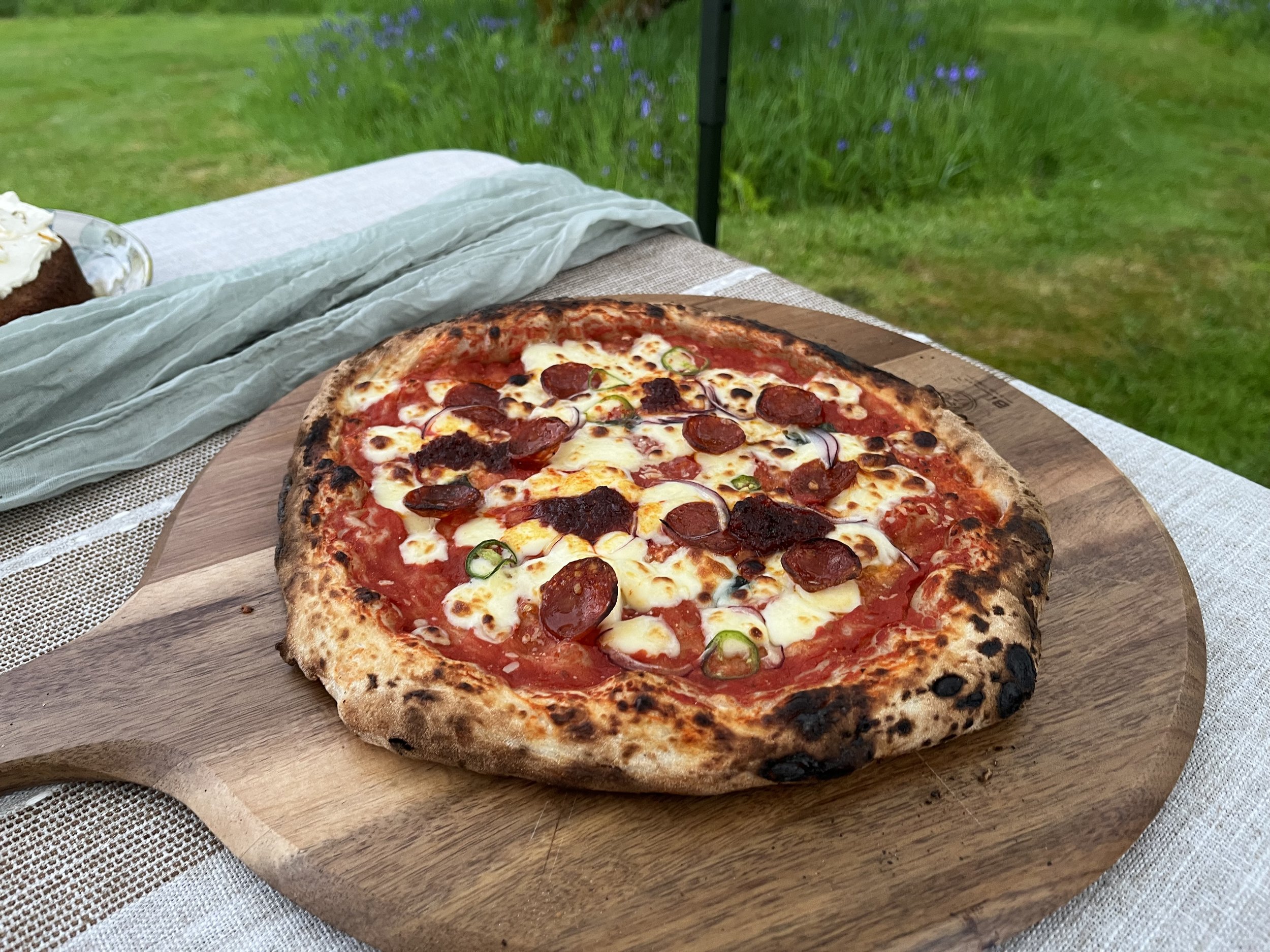 Close-up of a pizza with pepperoni, melted cheese, and green peppers on a wooden serving board, outdoors on a table with a grassy background.