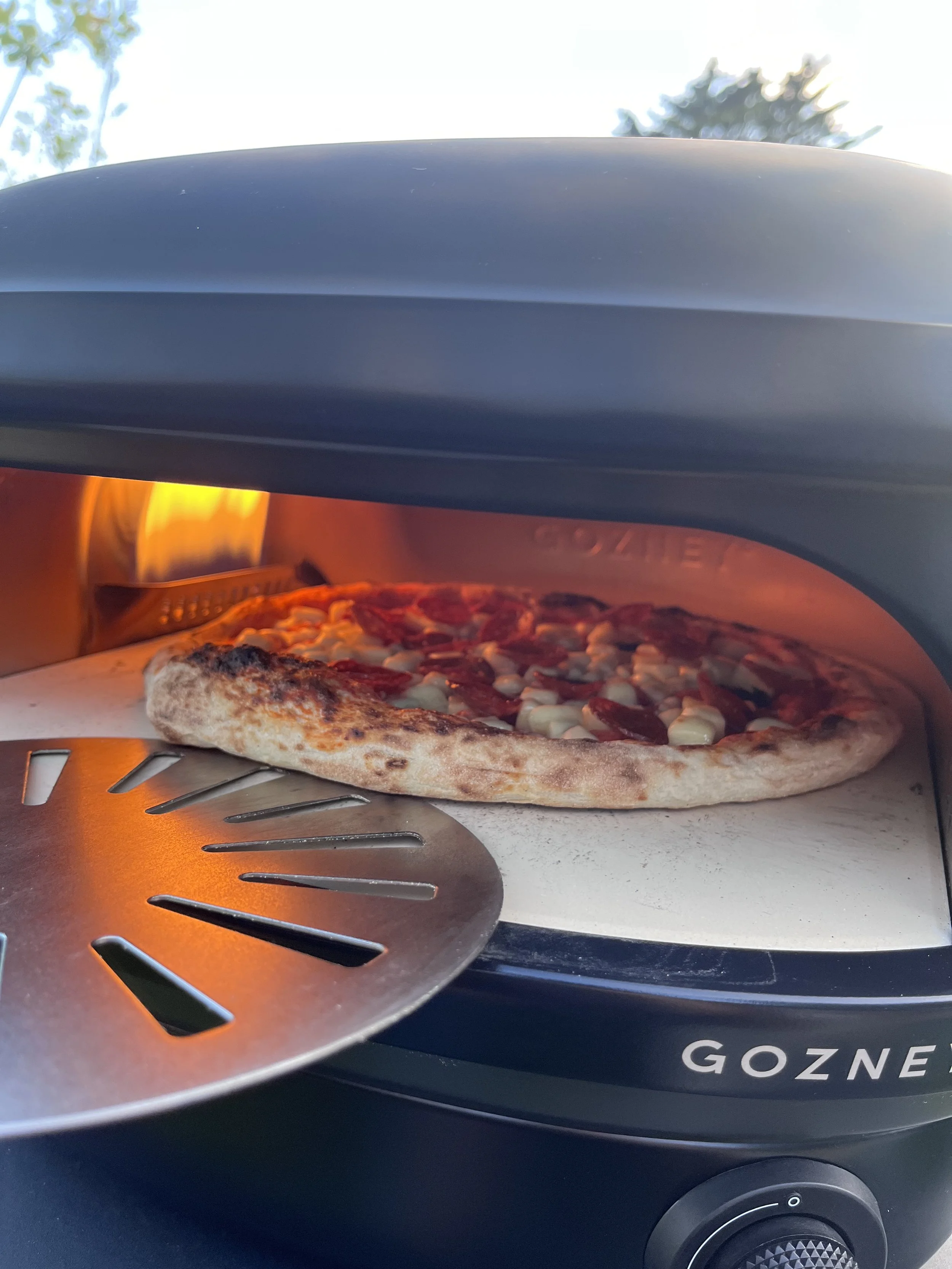A pizza with pepperoni and cheese baking inside a Gozney pizza oven. The oven's door is slightly open, revealing the glowing heat inside.