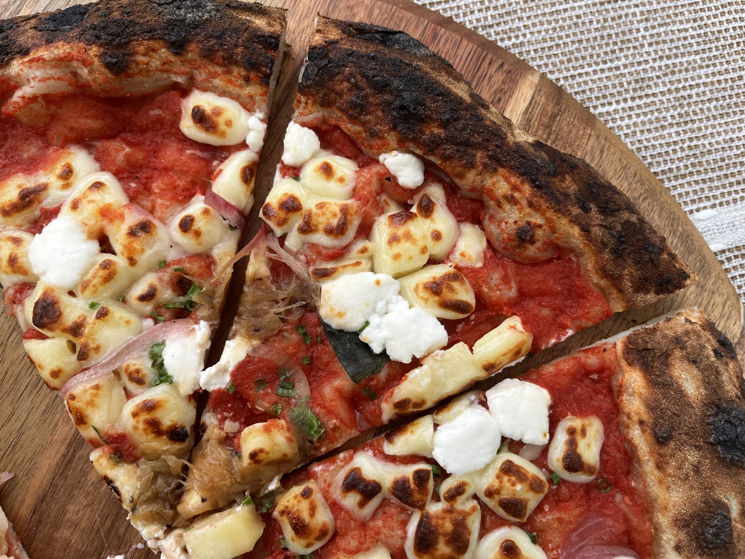 Close-up of several slices of pizza with various toppings on a round wooden board. The pizza has a charred crust and is topped with cheese, tomato sauce, and herbs.