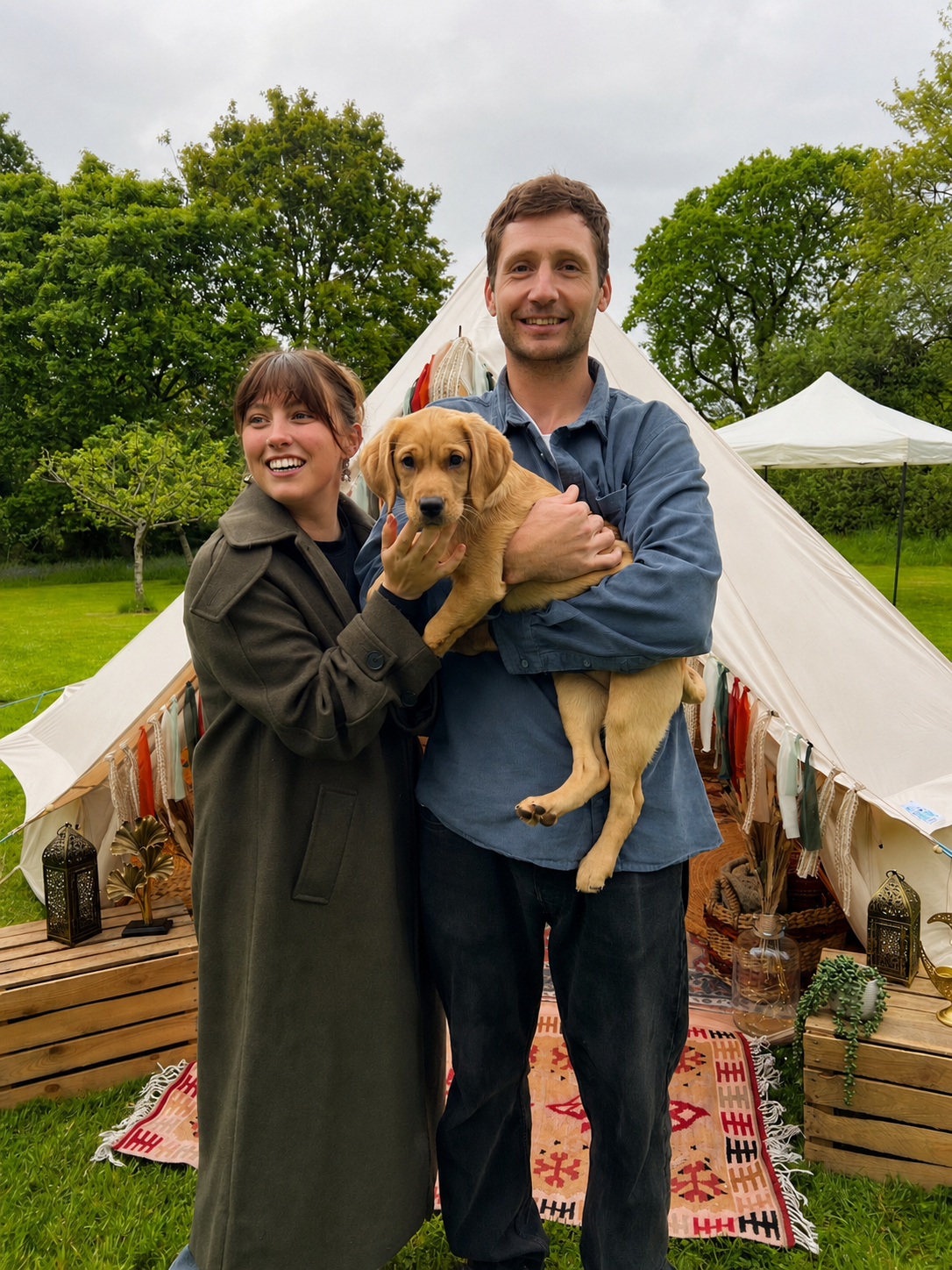 A happy man and woman are outdoors in front of a large white tent, holding a puppy. The man is holding the puppy, and the woman is smiling at him. There are trees and a grassy area in the background, and various decorative items are arranged on wooden palettes nearby.