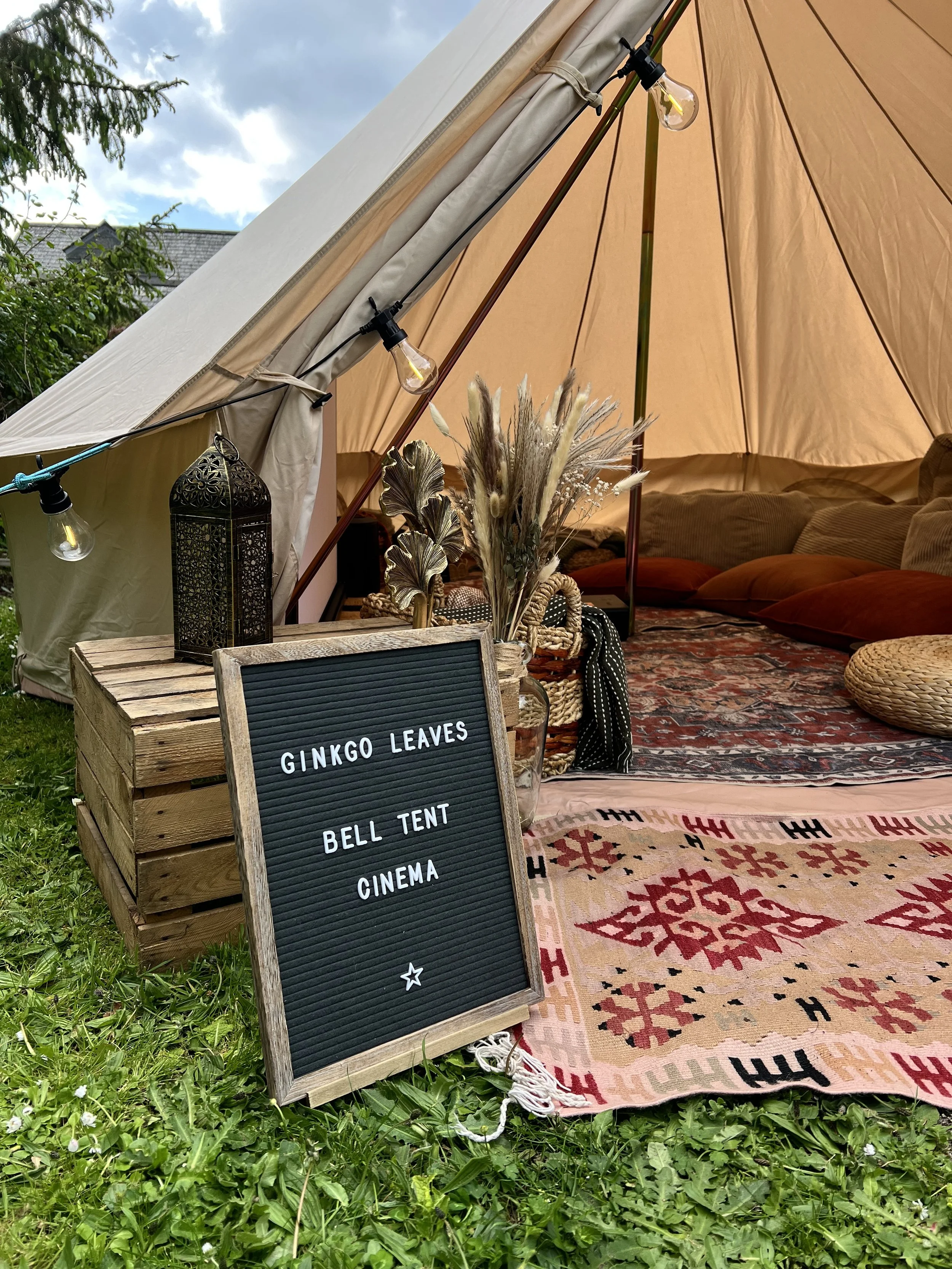 Outdoor tent setup with string lights, a decorative lantern, a vase with dried flowers, and a patterned rug. A sign with white text lists activities like Ginkgo Leaves, Bell Tent, and Cinema.