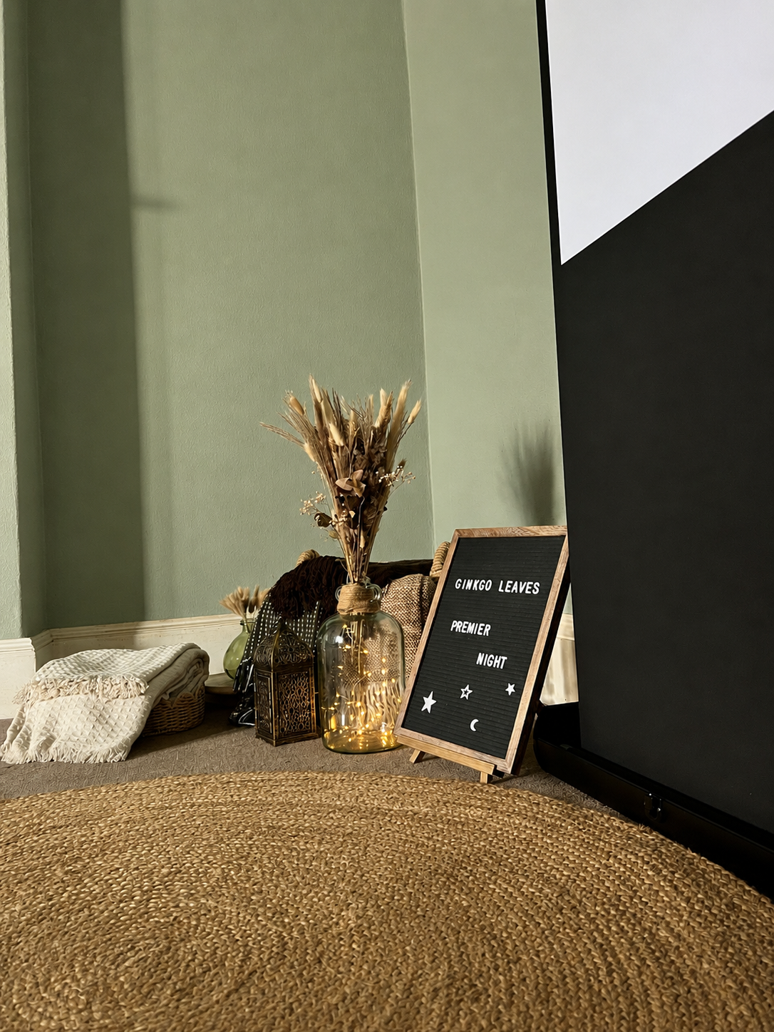 Decorative display with dried plants in a glass jar, a small lantern, and a black letter board that reads "Ginkgo Leaves Premier Night" with moon and star symbols, in a cozy, decorated corner of a room.