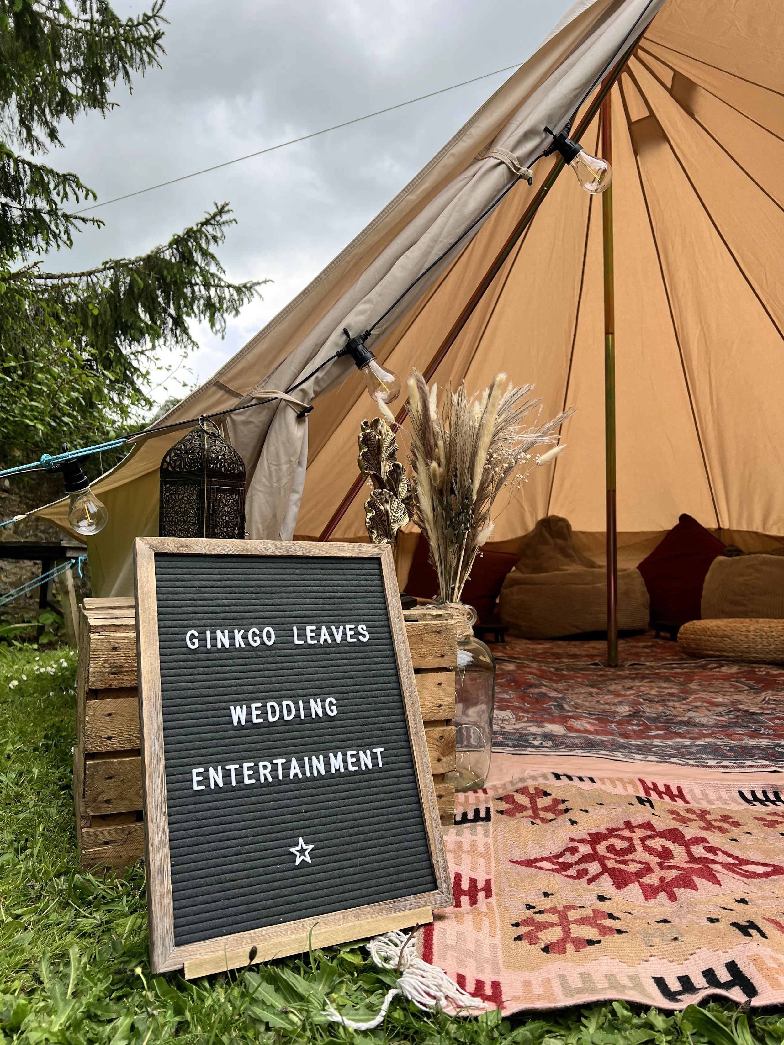 A wedding entertainment setup outdoors featuring a black felt letter board, a colourful patterned rug, and a bell tent decorated with plants and string lights, with green trees and a cloudy sky in the background.