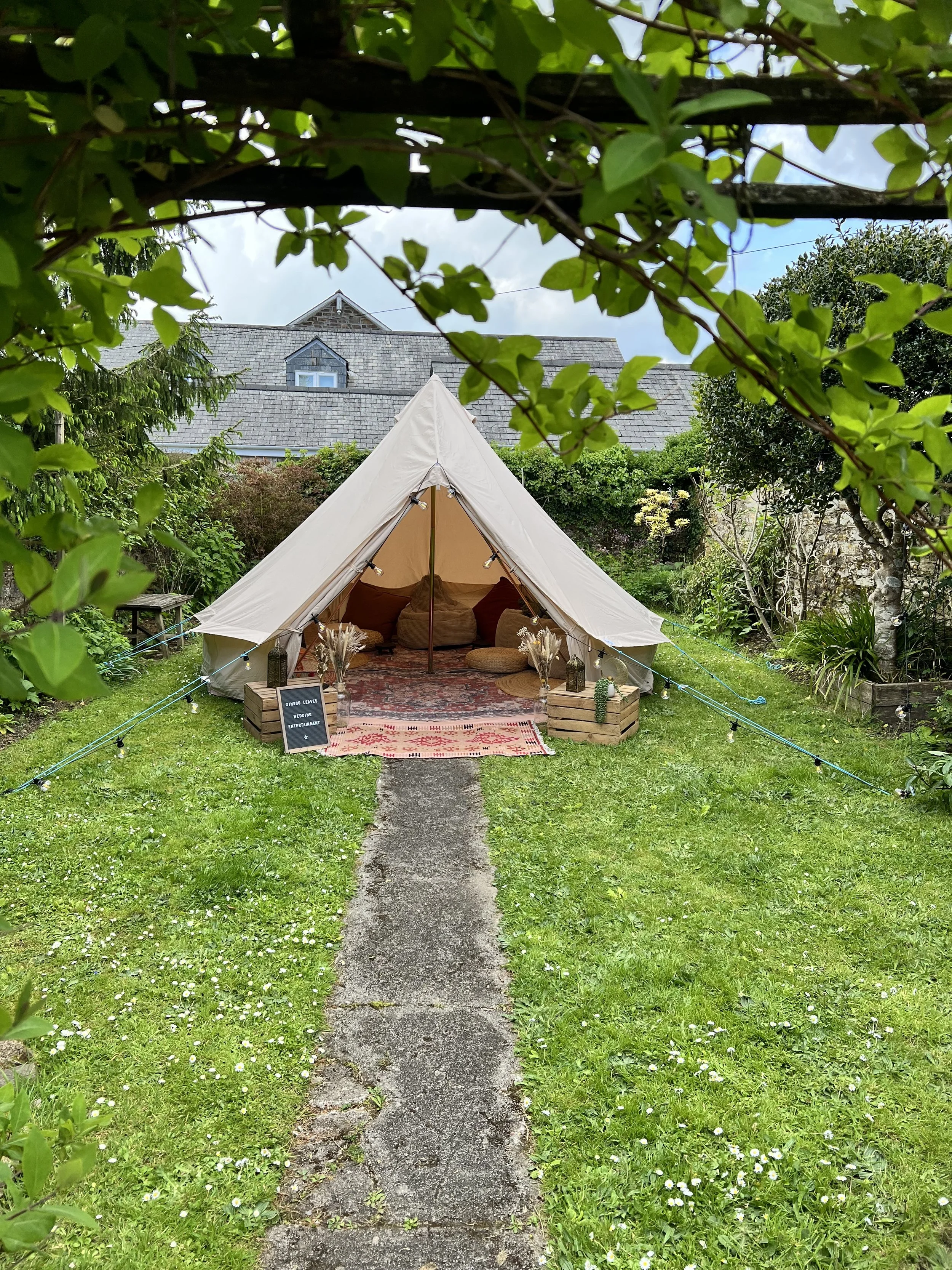 A garden tent set up with a pathway leading to it, surrounded by green grass, small white flowers, and lush green trees and shrubs, with a house roof in the background.