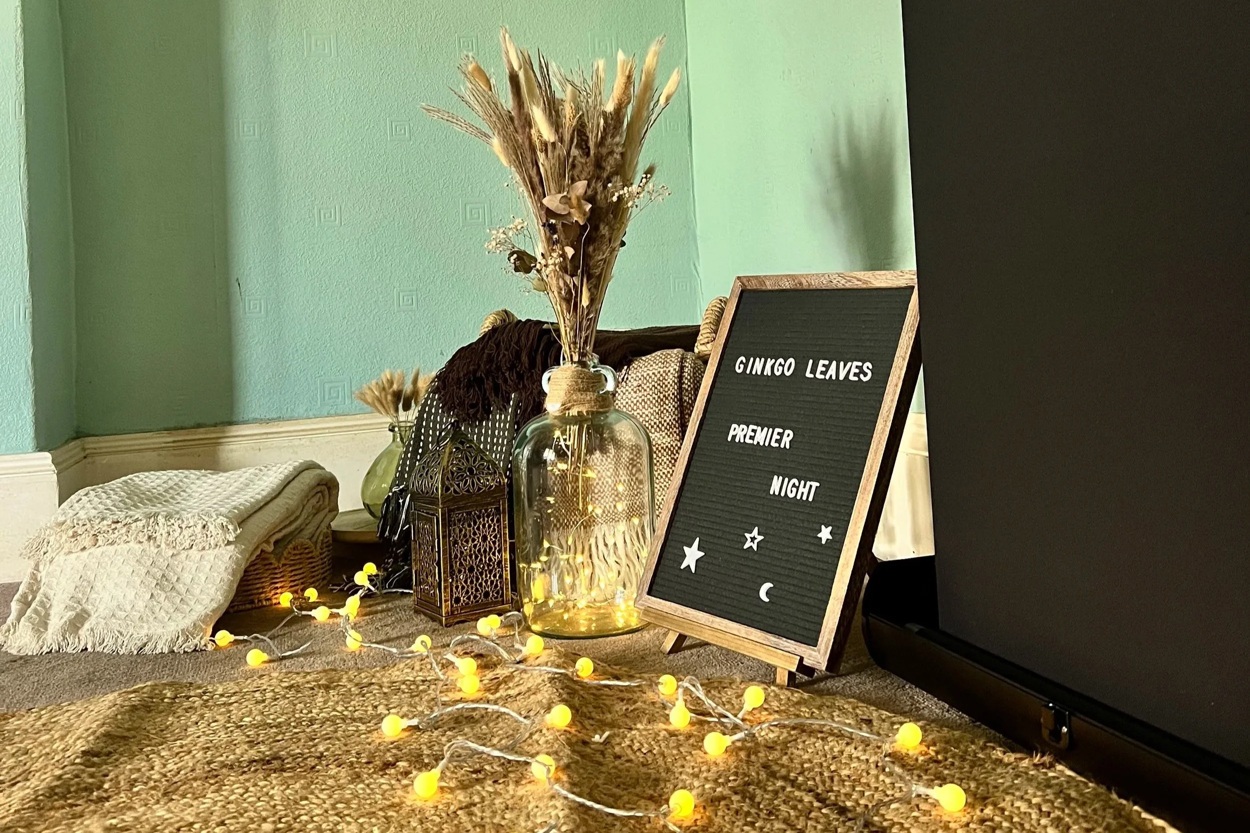 Decorative display with a black felt letter board with white text, dried flowers, fairy lights, and woven baskets on a plush carpet in a room with a light green wall.