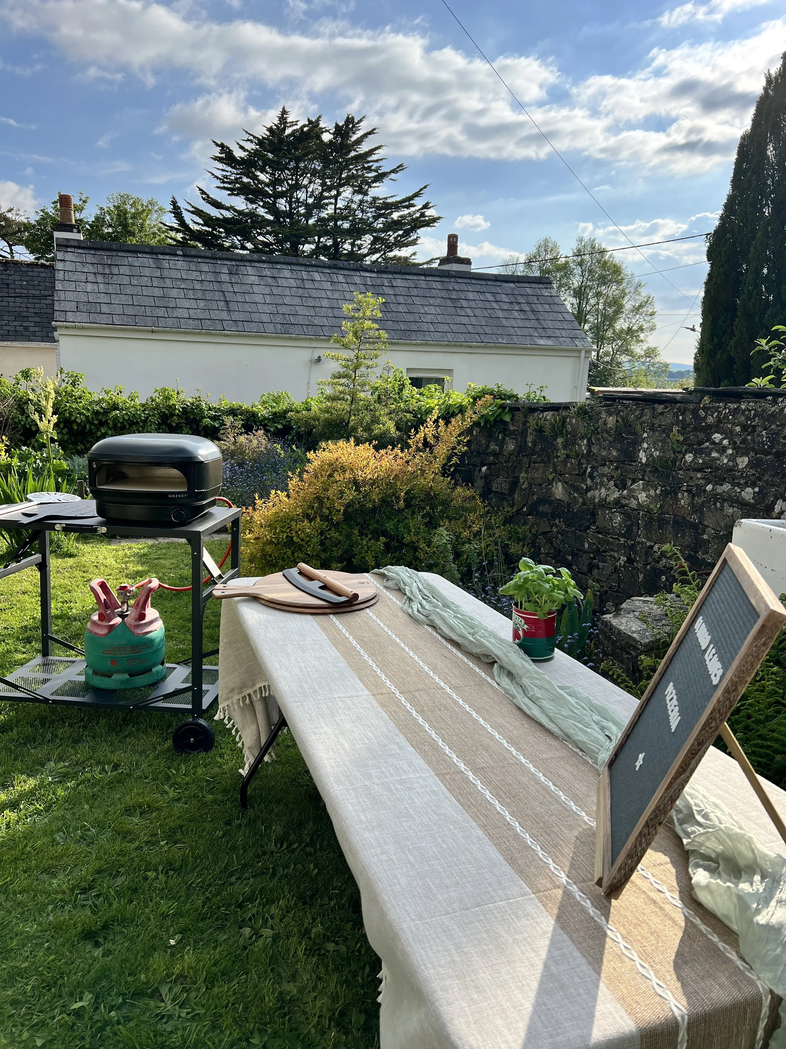 A backyard setting with a long table covered by a beige and white tablecloth, surrounded by various gardening tools, a small grill, a propane tank, and a chalkboard sign with writing. In the background, there are plants, a stone wall, trees, and houses under a partly cloudy sky.
