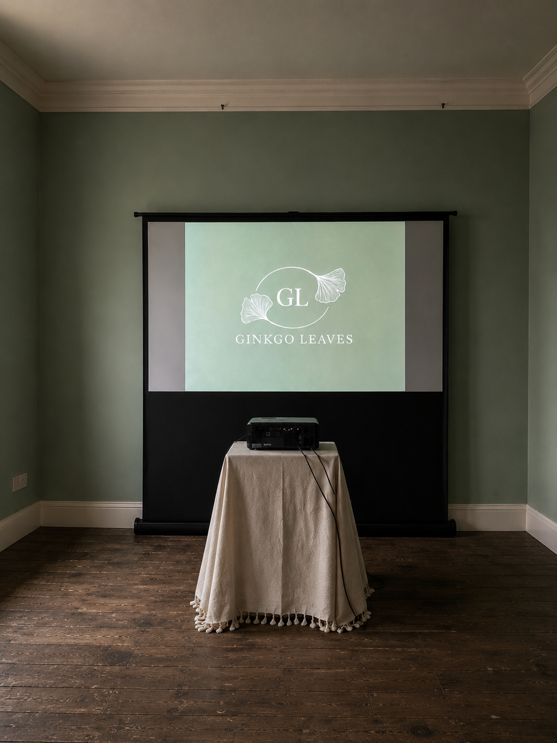 A presentation setup with a projector on a table covered with a white cloth, projecting a slide for Ginkgo Leaves featuring the initials "GL" and ginkgo leaf illustrations inside a circle, against a green wall.