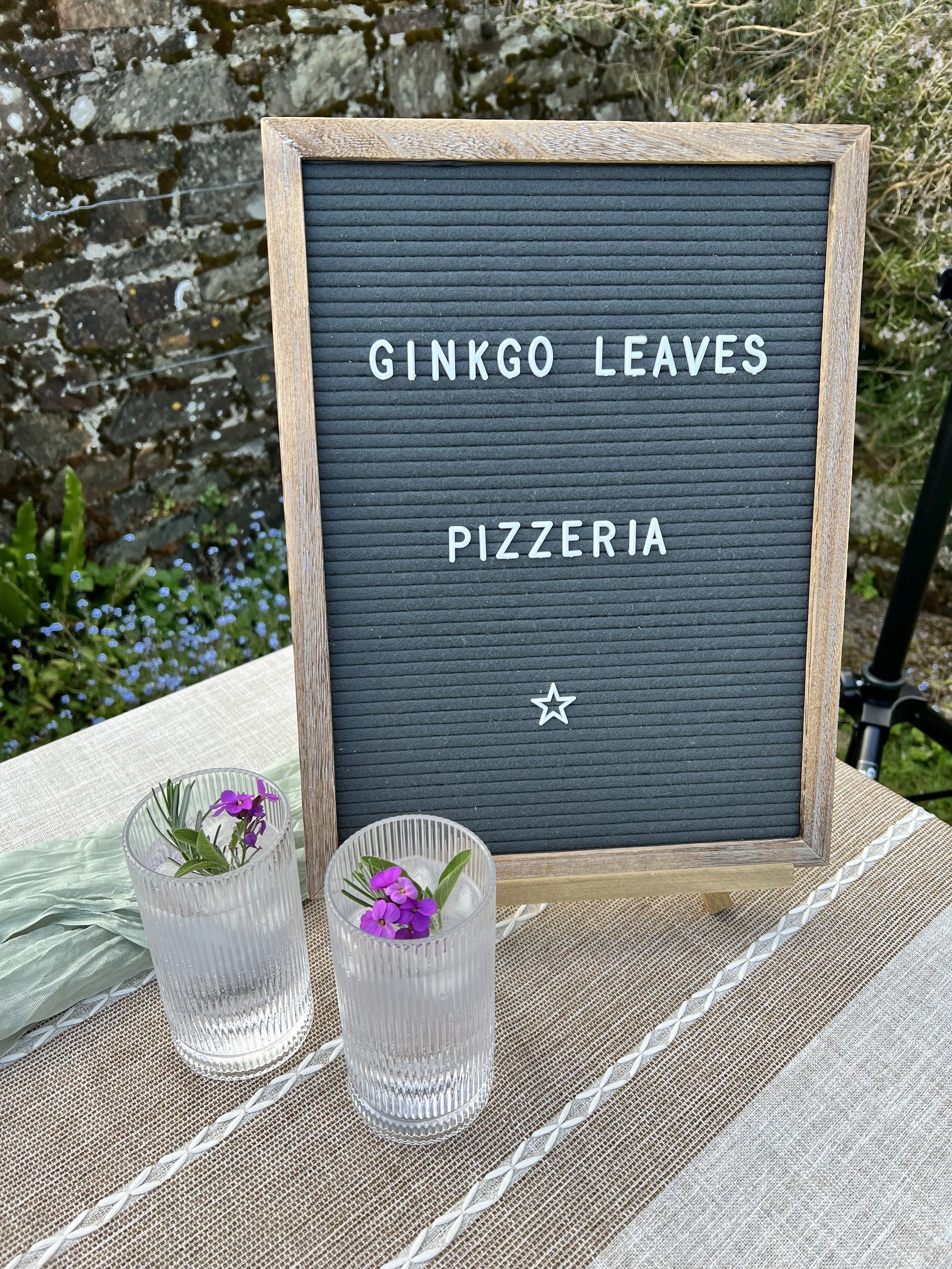 A black letter board with a wooden frame displaying the words "Ginkgo Leaves Pizzeria" and a small star icon, set on a table decorated with two small glass vases of purple flowers, with greenery and a stone wall in the background.