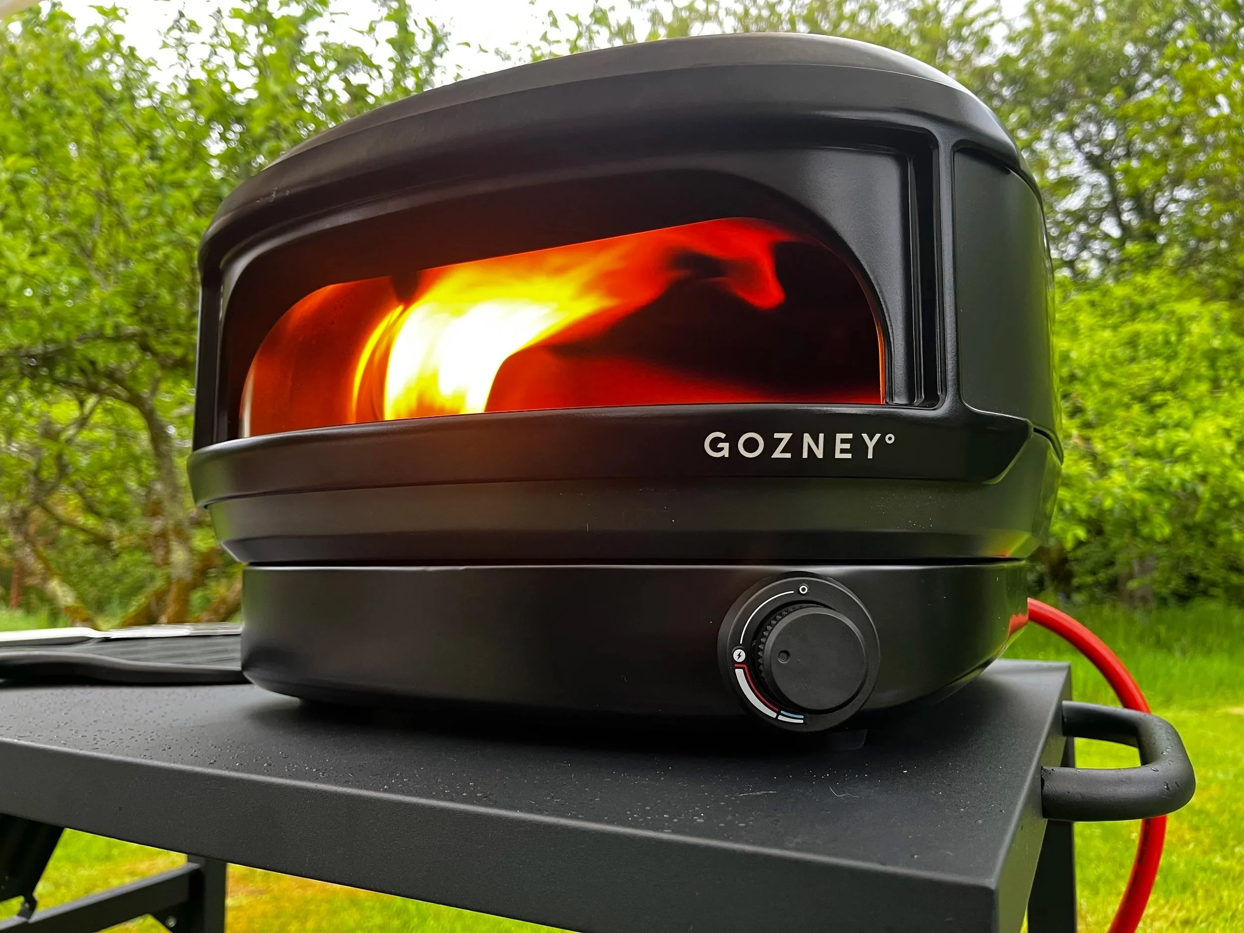 Close-up of a black portable infrared outdoor heater with a glowing heating element inside, placed on a black table outdoors with green trees in the background.