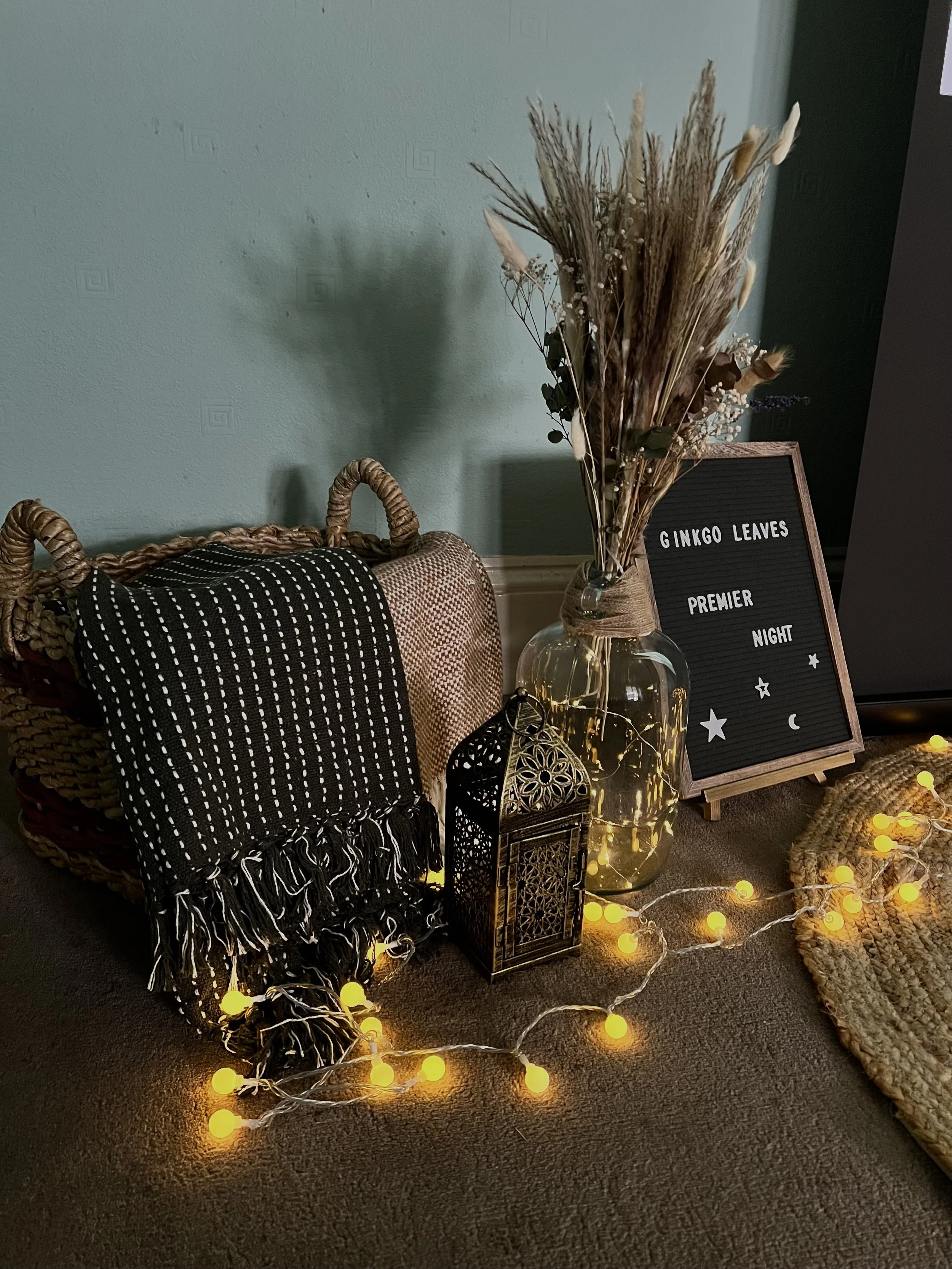 A cozy arrangement with a wicker basket, a black and white fringed throw, a glass vase with dried flowers, a letter board with celestial words, and yellow string lights on a carpeted floor.
