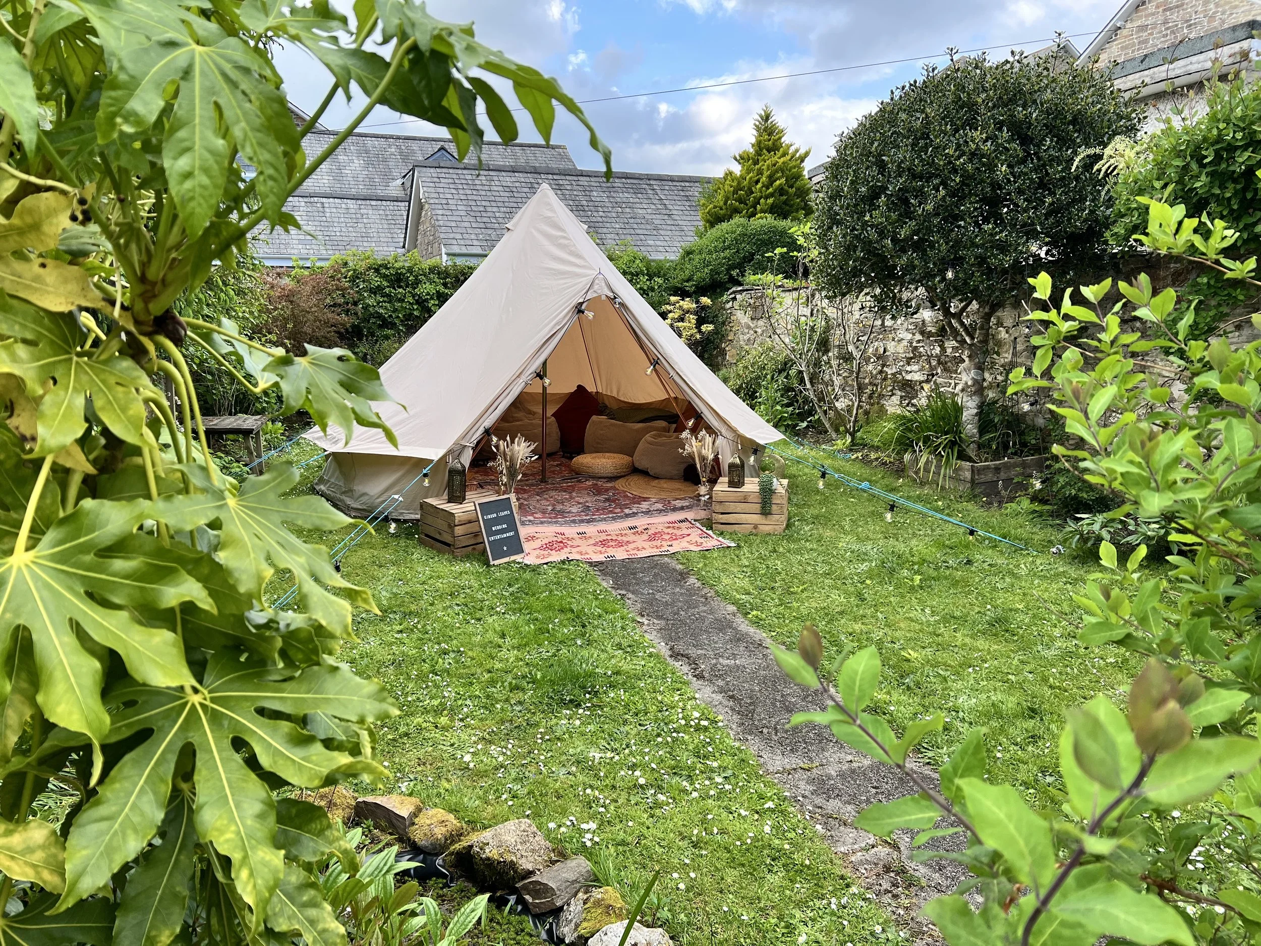 A backyard scene featuring a triangular off-white tent with cushions inside, a colorful rug at the entrance, surrounded by green trees, shrubs, and plants, with a stone and brick wall behind. A small signboard and decorative lanterns are placed near the tent.
