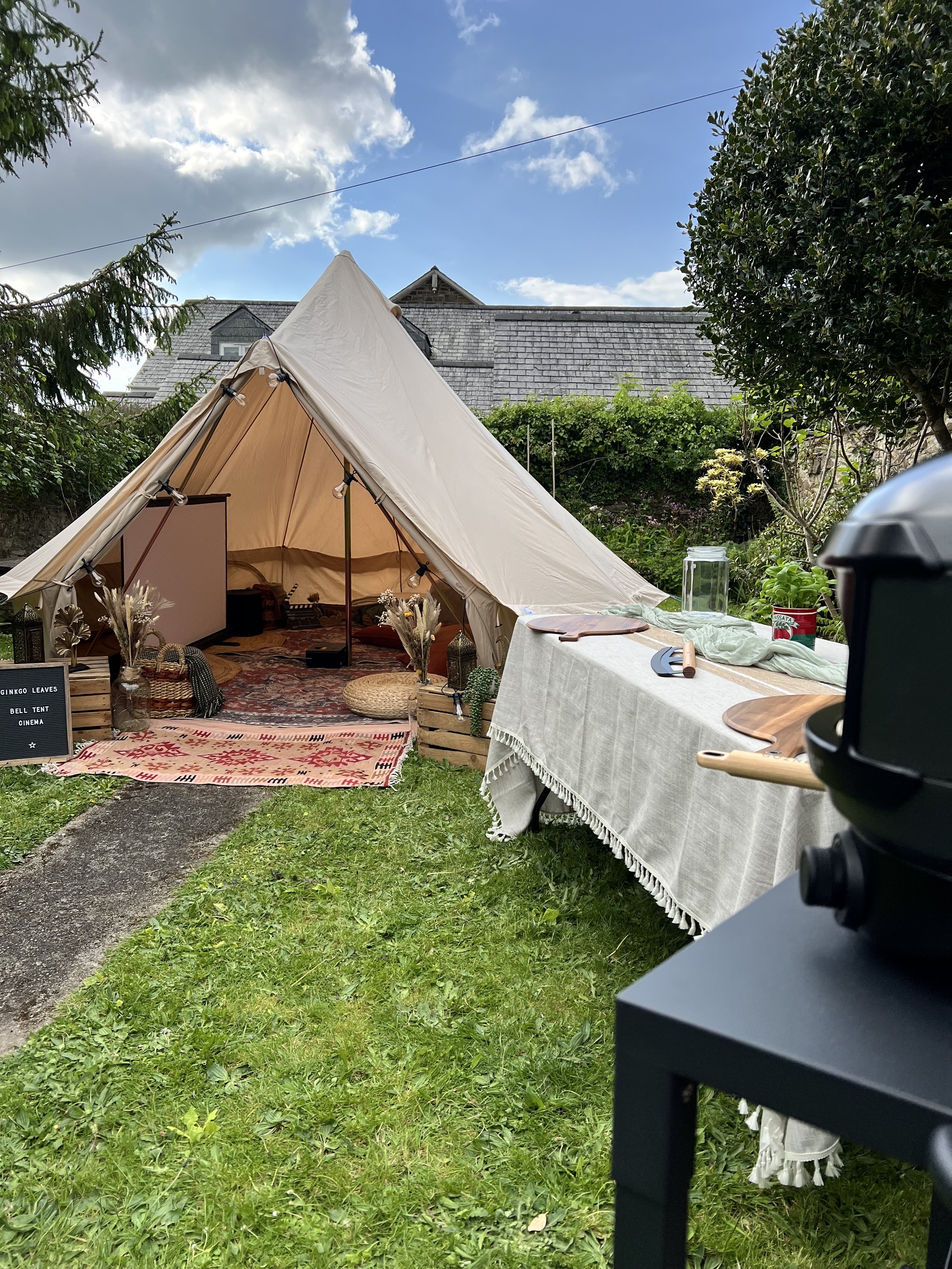 A cream-colored bell tent set up outdoors on grass with a blue sky and some clouds overhead. Surrounding the tent are trees and greenery. In front of the tent is a table covered with a white cloth, with various items including jars, greenery, and wooden utensils on it.