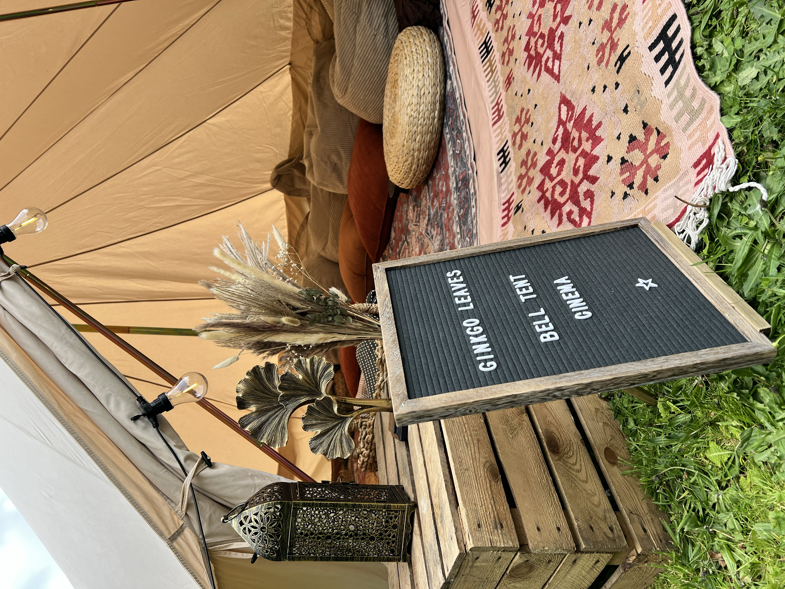 A cozy outdoor seating area with a beige beach tent, patterned rugs, throw pillows, and green plants. A wooden table holds decorative dried flowers and a signboard with text.