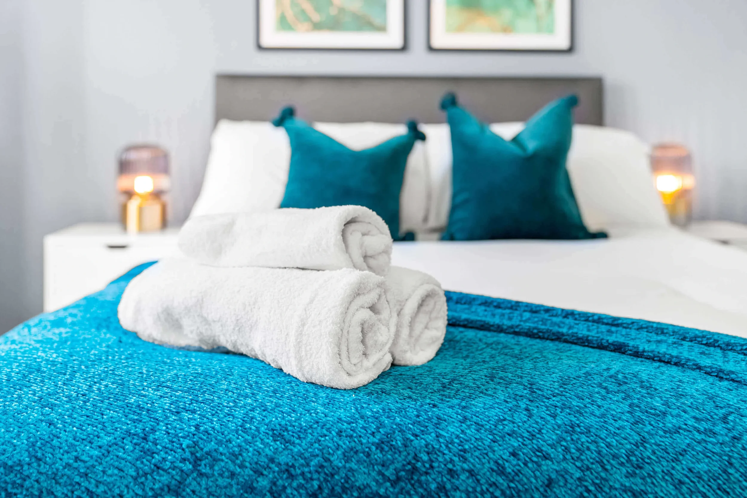 A neatly made bed with a white towel rolled on a blue textured blanket, two teal pillows, and two lamps on nightstands on either side of the bed, with framed artwork hanging on the wall above.